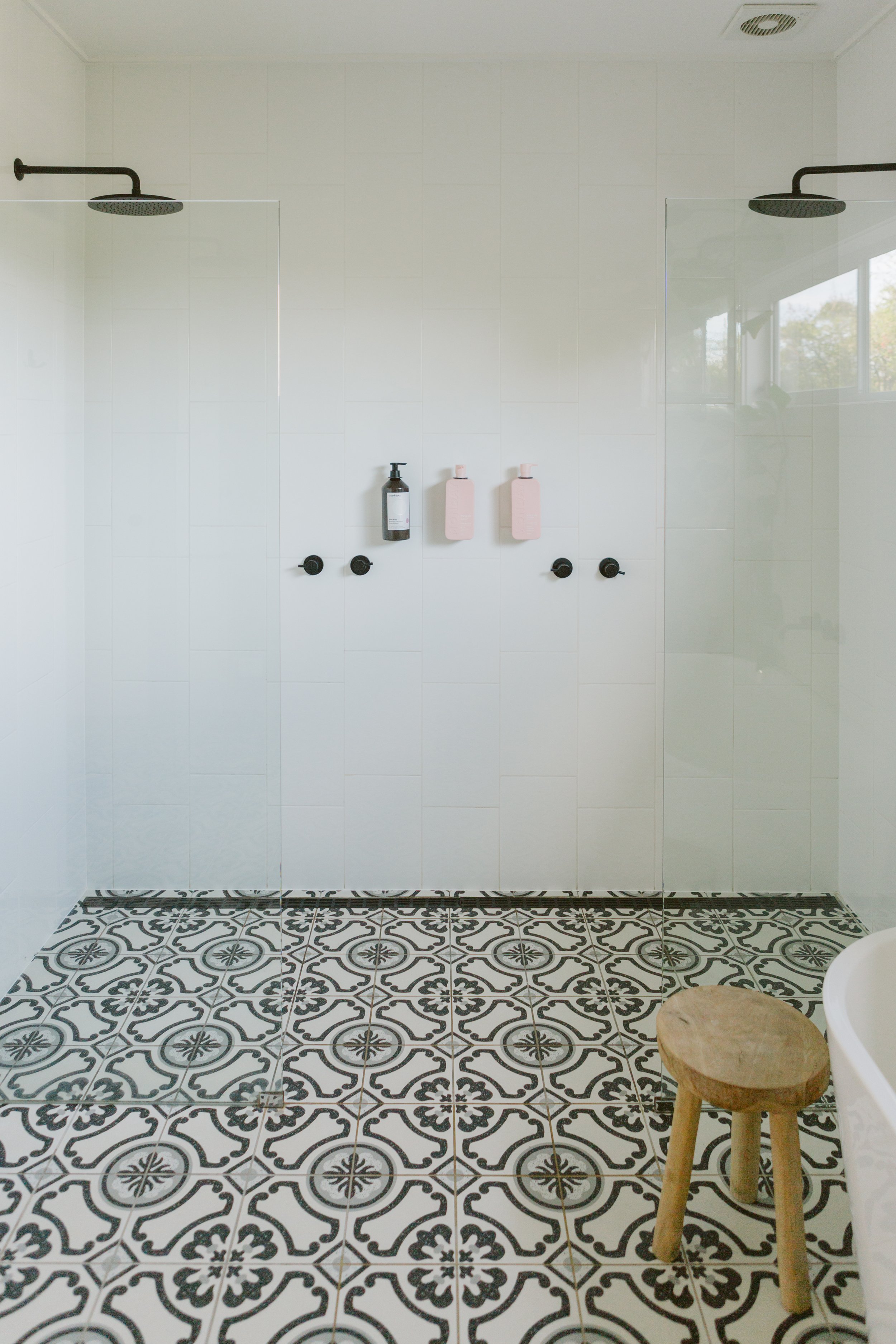 Modern bathroom with patterned black and white floor tiles, featuring a glass shower enclosure with black fixtures, three black wall hooks, and two pink soap dispensers, a wooden stool, and a window with natural light.
