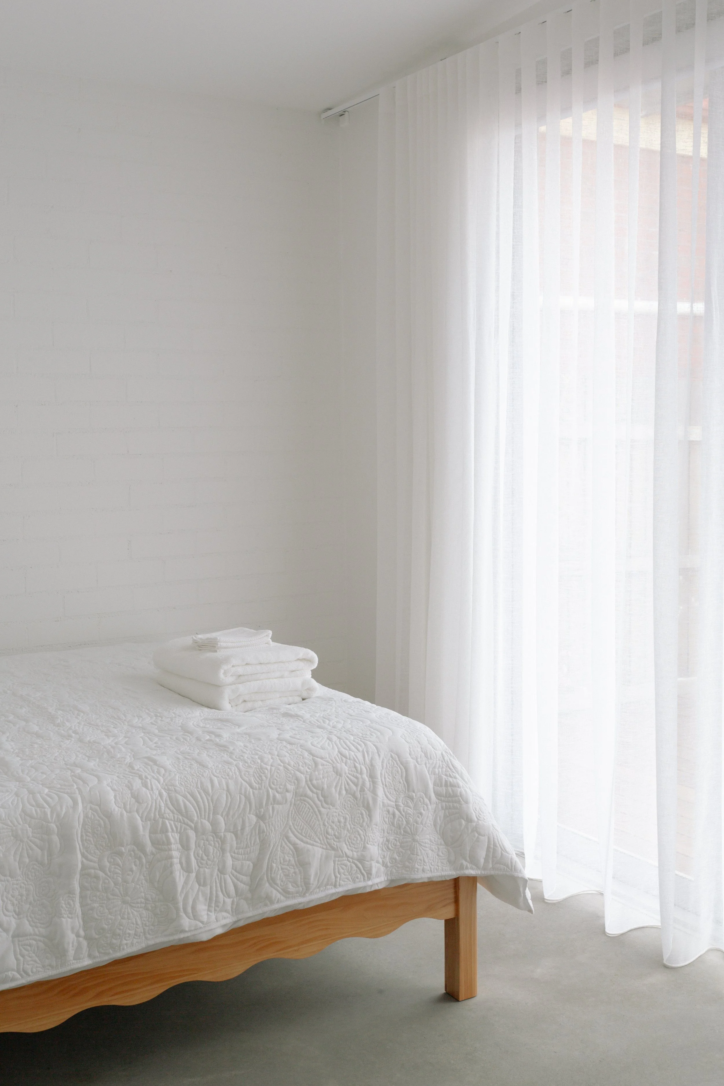 A minimalist bedroom with a neatly made bed covered in a white quilt, folded towels on top, and sheer white curtains letting in natural light.