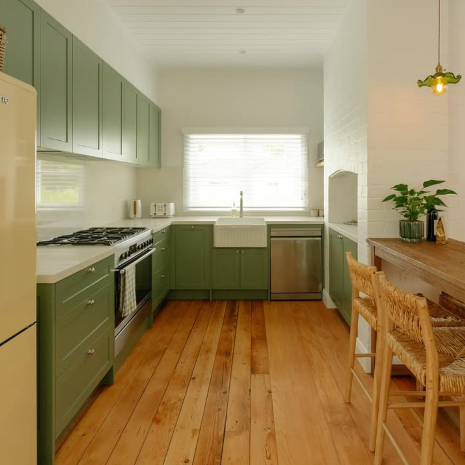 A cheerful kitchen with green cabinets, white countertops, a large window with blinds, and a wooden floor. Features include a stove, a dishwasher, a farmhouse sink, and a small dining area with wicker chairs and a wooden table with a potted plant.