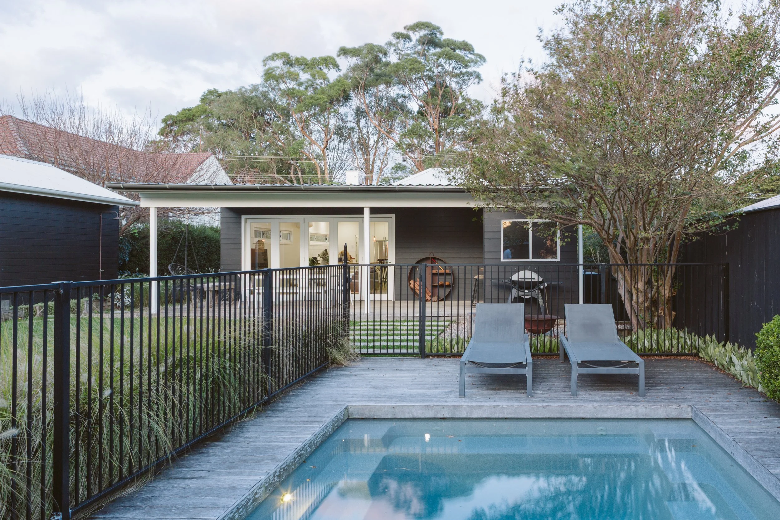 Backyard with swimming pool, two lounge chairs, black metal fence, trees, and modern house with glass sliding doors.