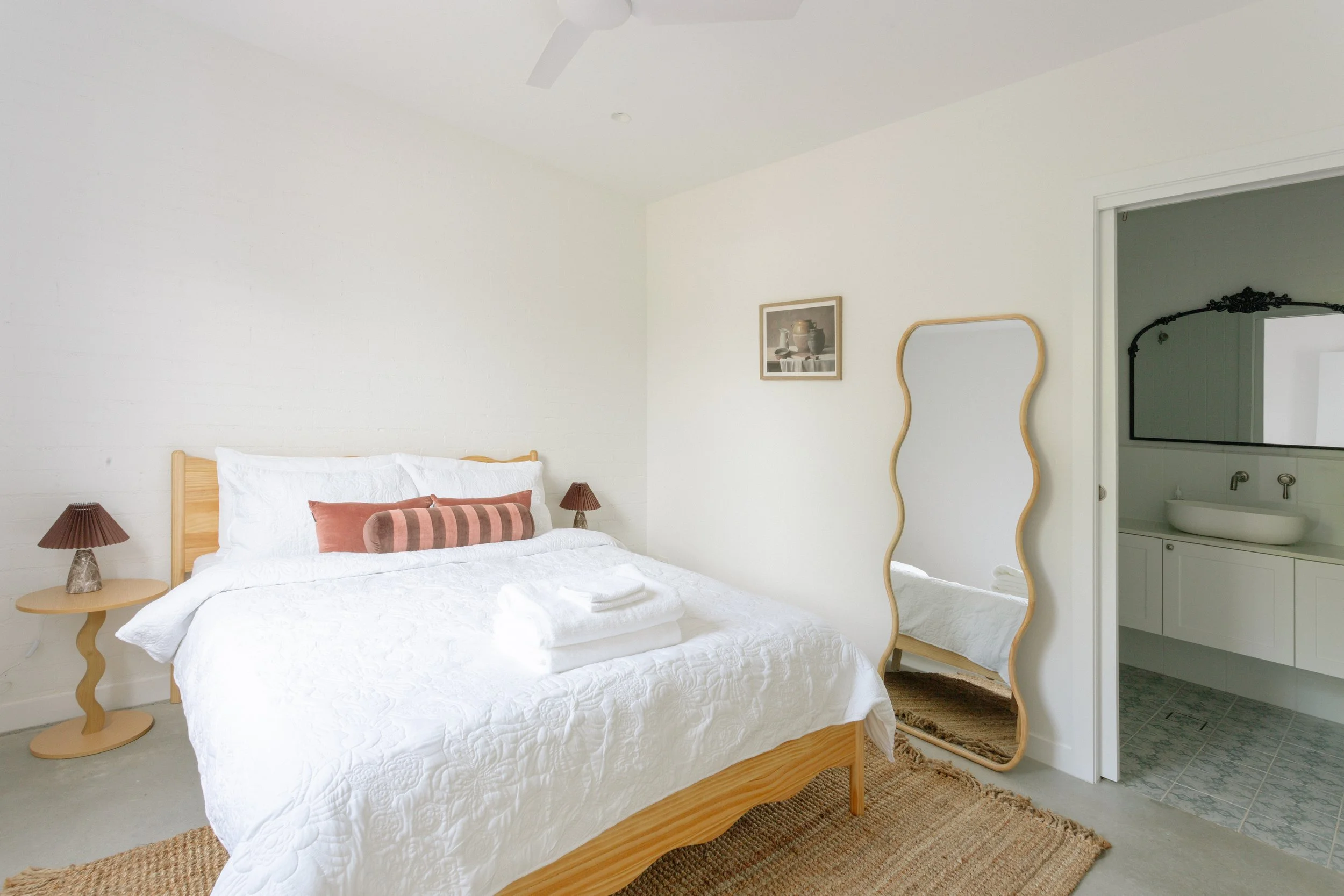 A bedroom with a wooden bed frame, white bedding, and pillows; two side tables with small lamps; a wavy full-length mirror; a small piece of art on the wall; an open door leading to a bathroom with white vanity and black framed mirror.