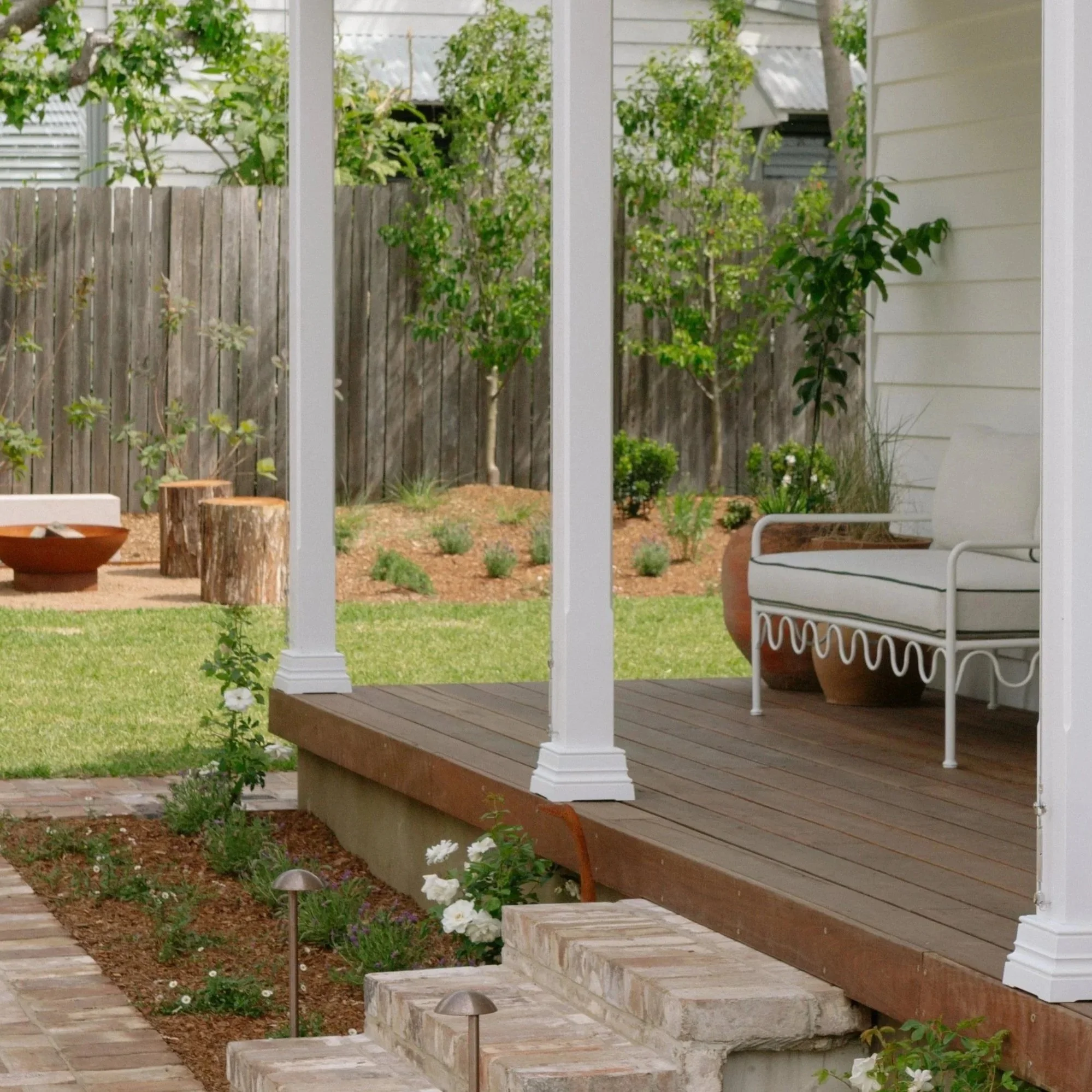 A cozy backyard porch with white framing, a more modern white outdoor sofa, potted plants, and a view of a backyard with a grass lawn, trees, small bushes, and a wooden fence.