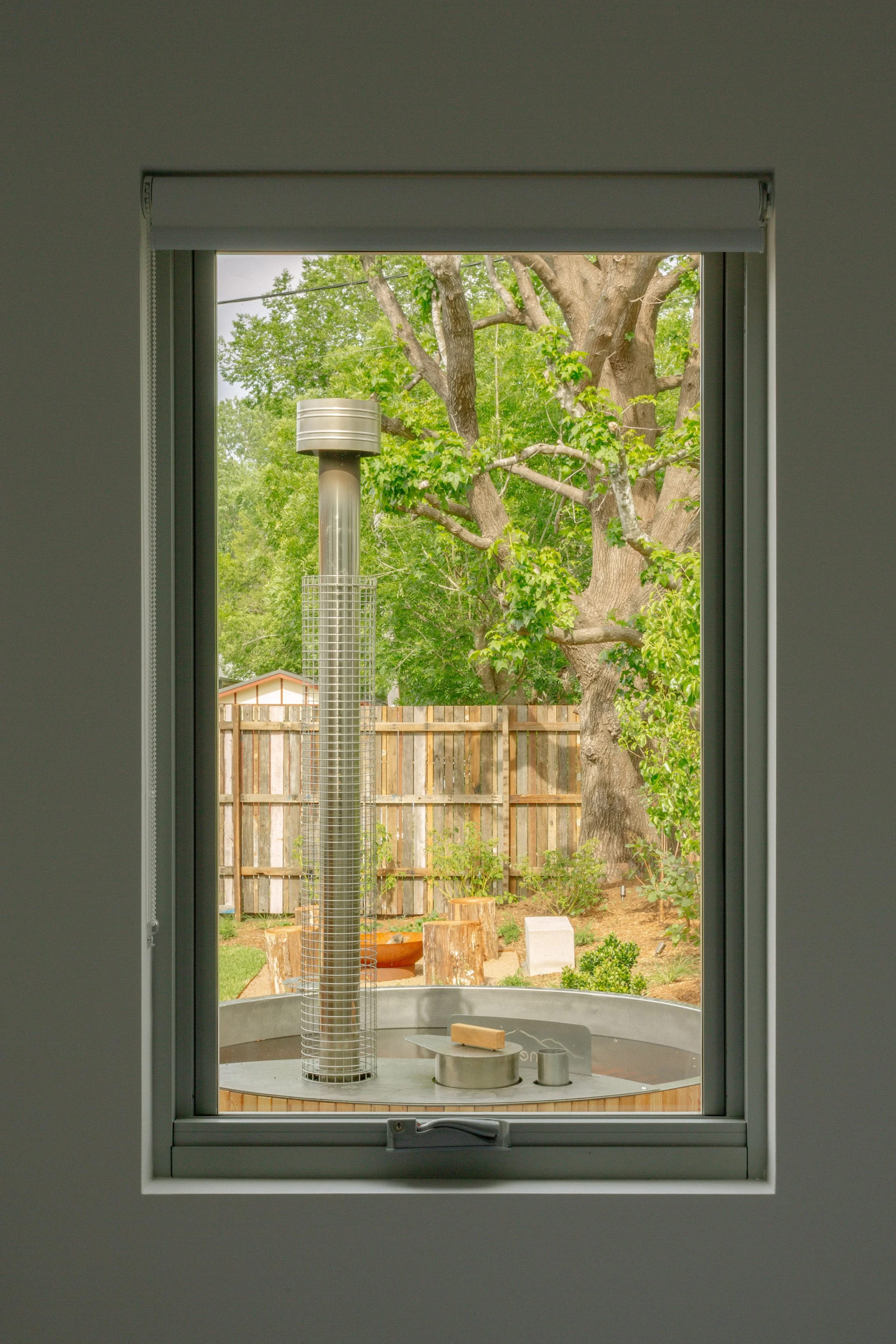View through a window overlooking a backyard with a large tree, wooden fence, and some garden decor, including wood stumps and small plants.