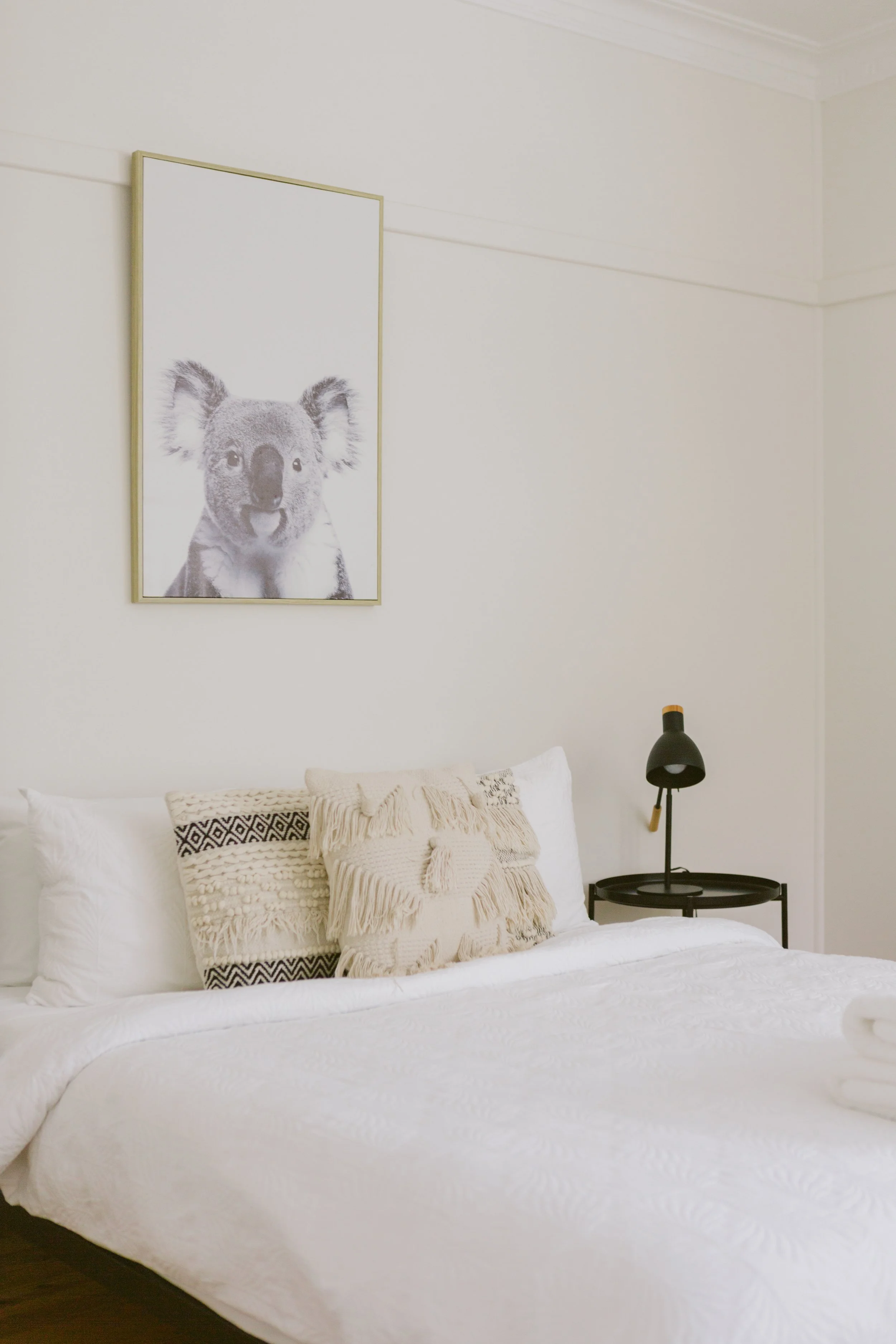 A cozy bedroom with a white bed, decorative pillows, a black bedside table, a black adjustable desk lamp, and a framed black-and-white art print of a koala bear on the wall.
