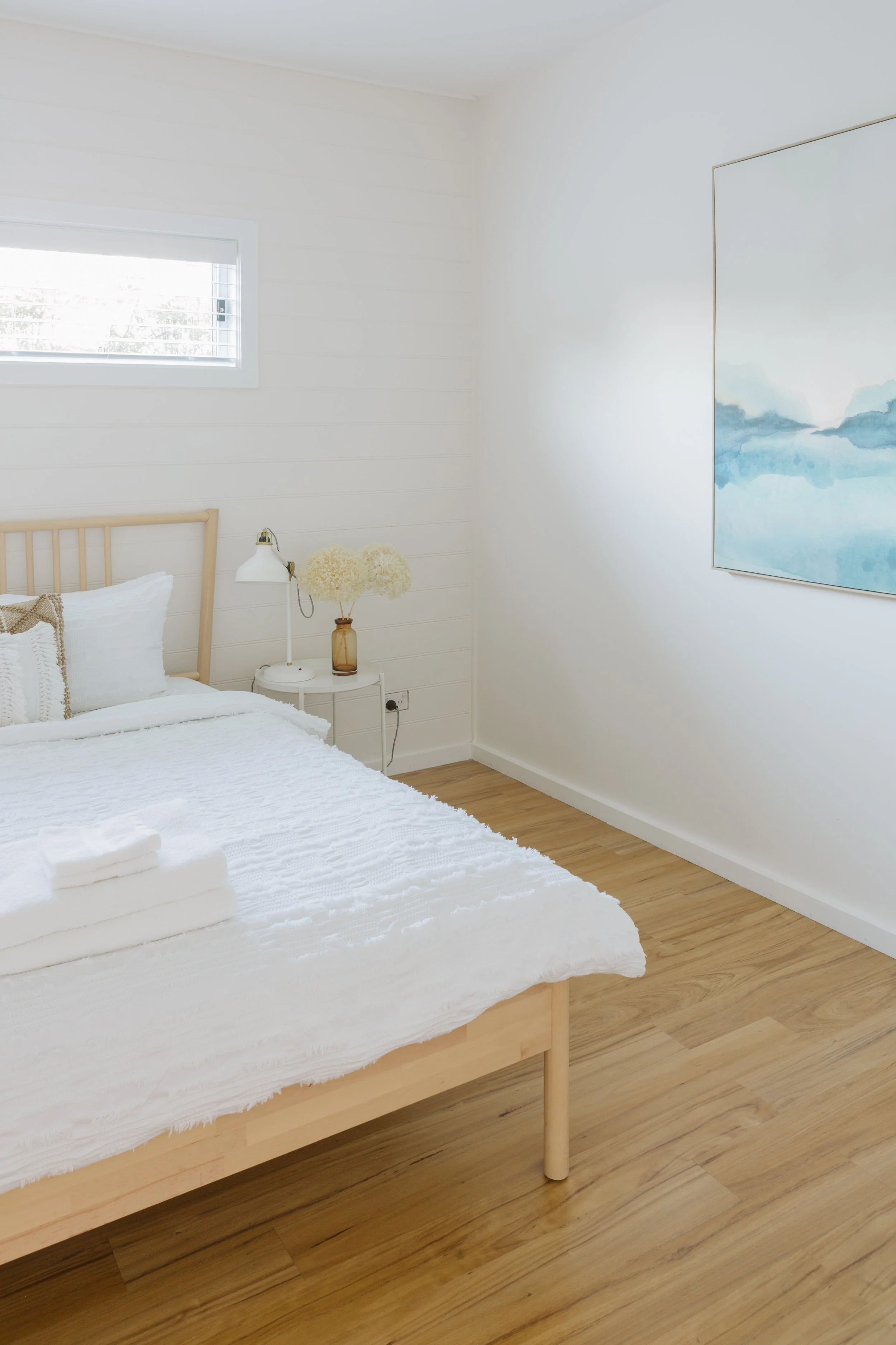 A minimalistic bedroom with white walls, a wooden bed with white bedding, a small bedside table with a lamp and a vase with flowers, and a large blue watercolor painting on the wall.