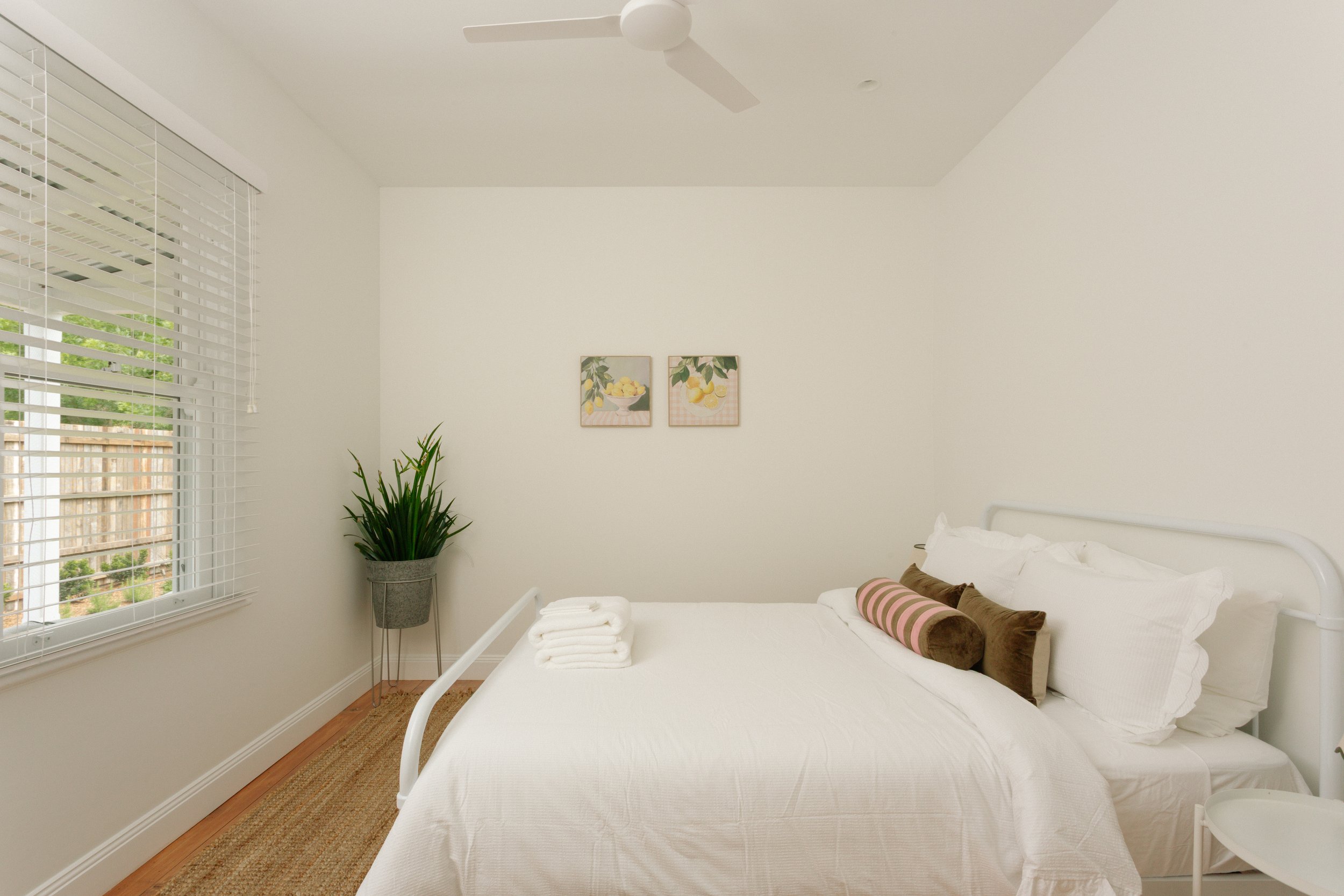 A bright bedroom with white walls, a window with white blinds, a white bed with multiple pillows, a rolled brown and pink bolster pillow, a potted plant in a standing gray pot, and two framed botanical pictures on the wall.