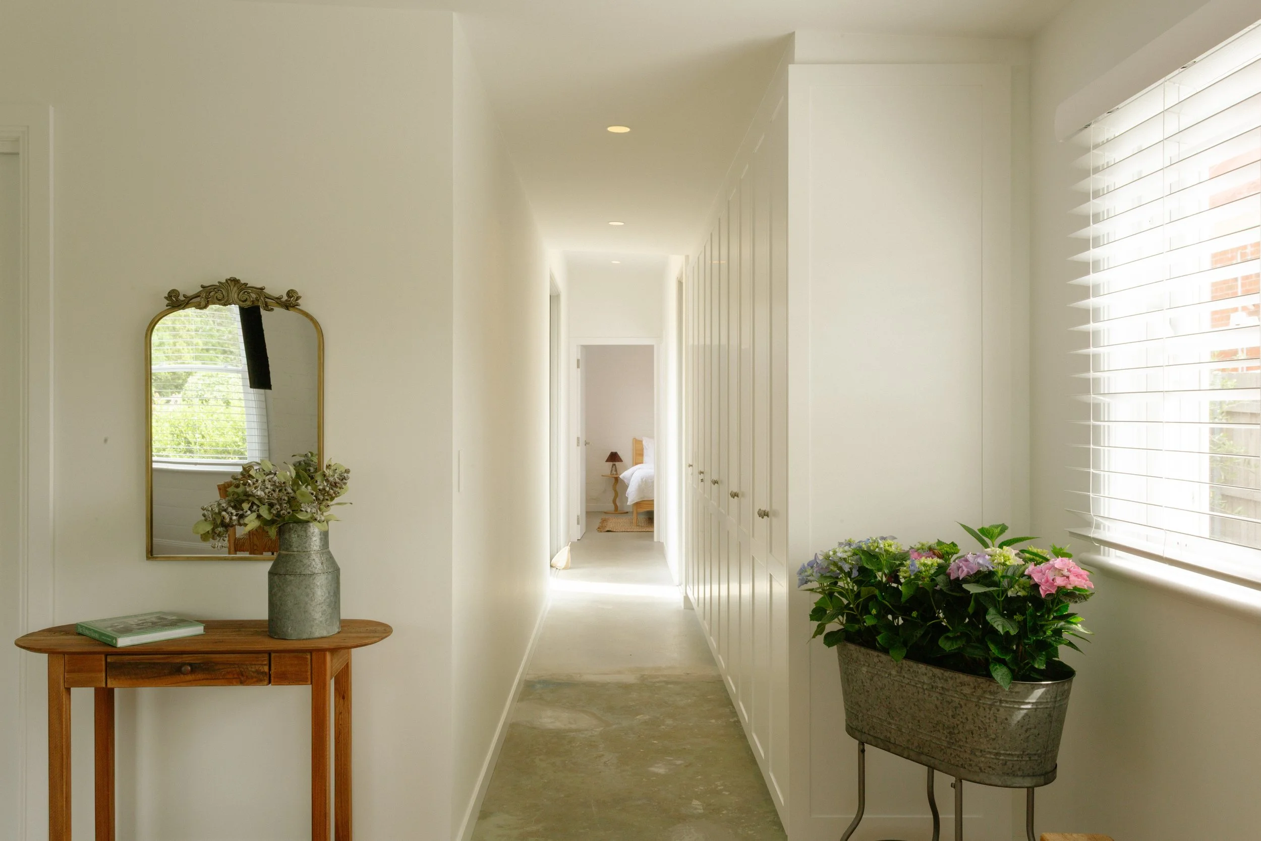 Bright hallway with white walls, wooden console table with mirror and vase on the left, flowering plant in a metal planter on the right, and a bedroom visible at the end of the hallway.