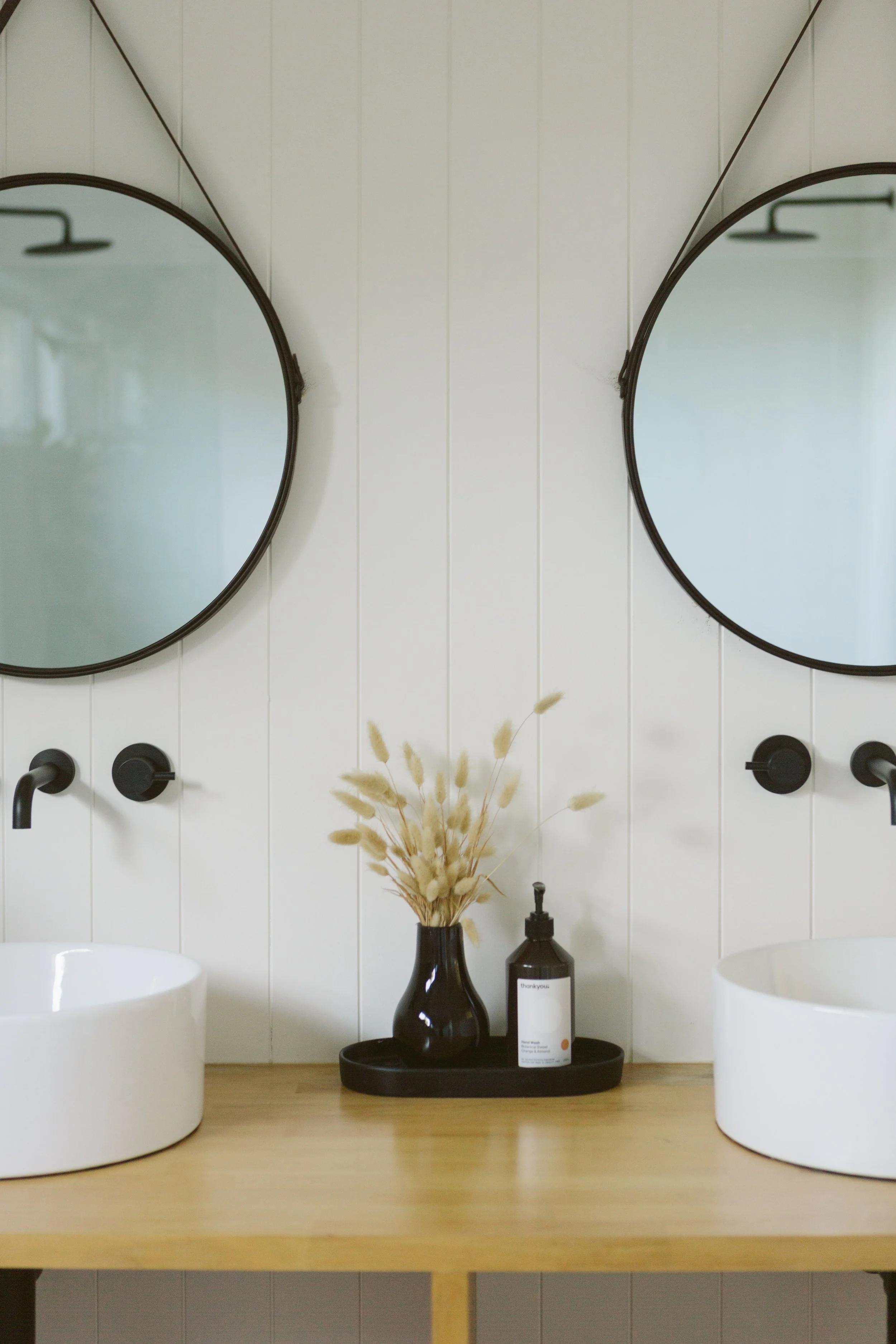 Bathroom double vanity with two round mirrors, white vessel sinks, black wall-mounted faucets, a black tray with a vase of dried plants, and a soap dispenser on a wooden countertop.