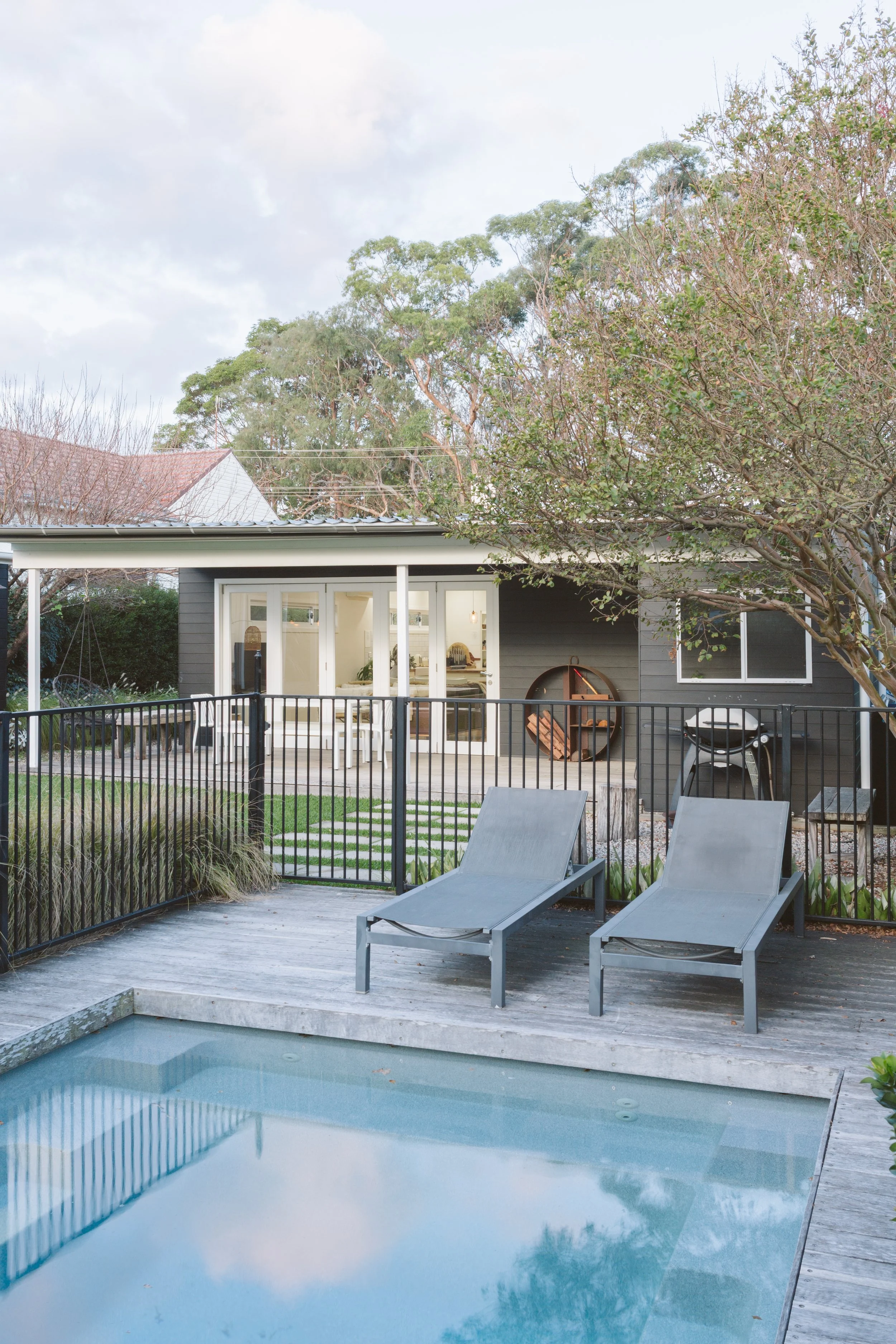 A backyard with a swimming pool, two lounge chairs, a fence, and a house with large glass doors and windows, surrounded by trees.