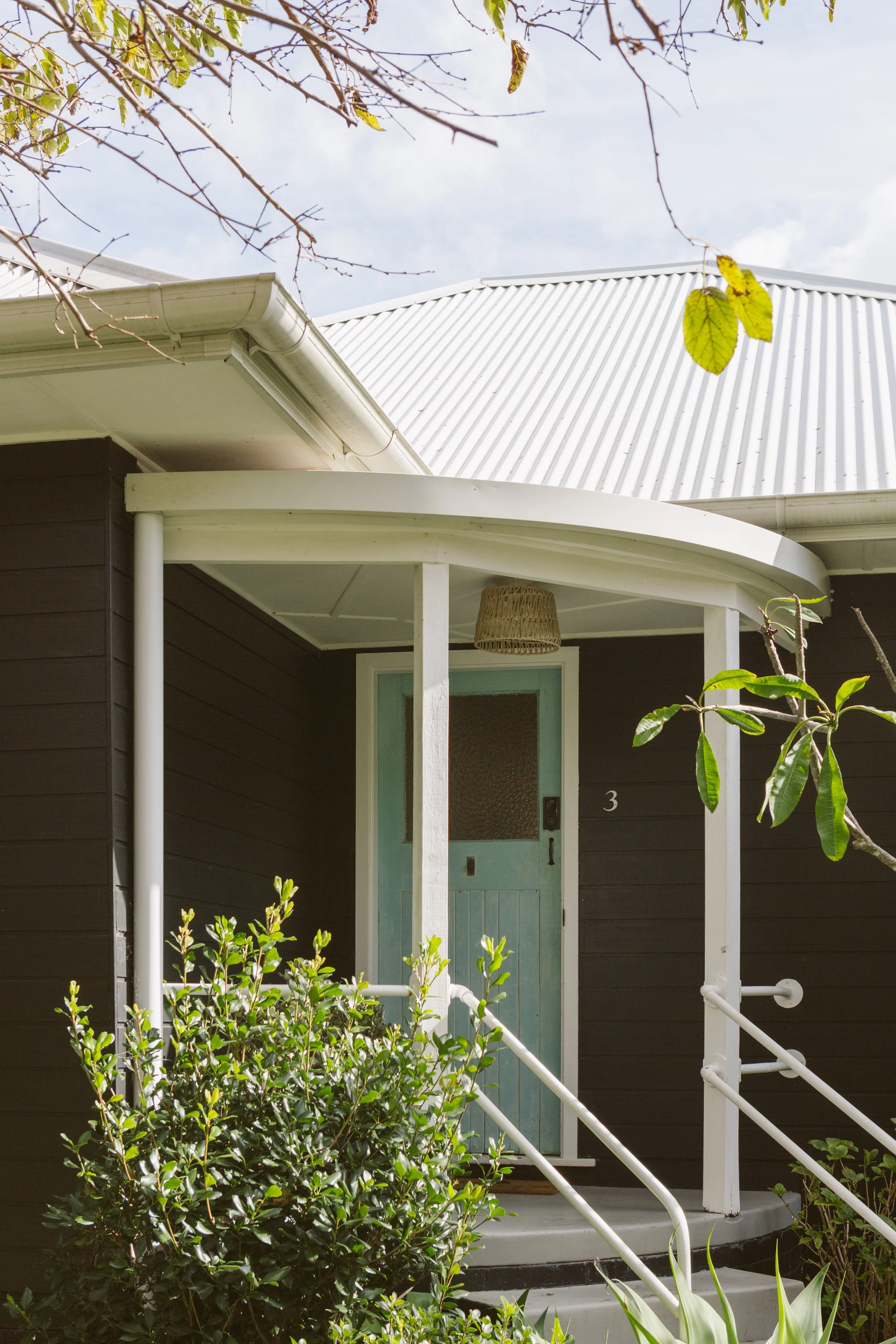 Front porch of a dark-colored house with a light blue door, outdoor greenery, and a white metal handrail, under a cloudy sky.