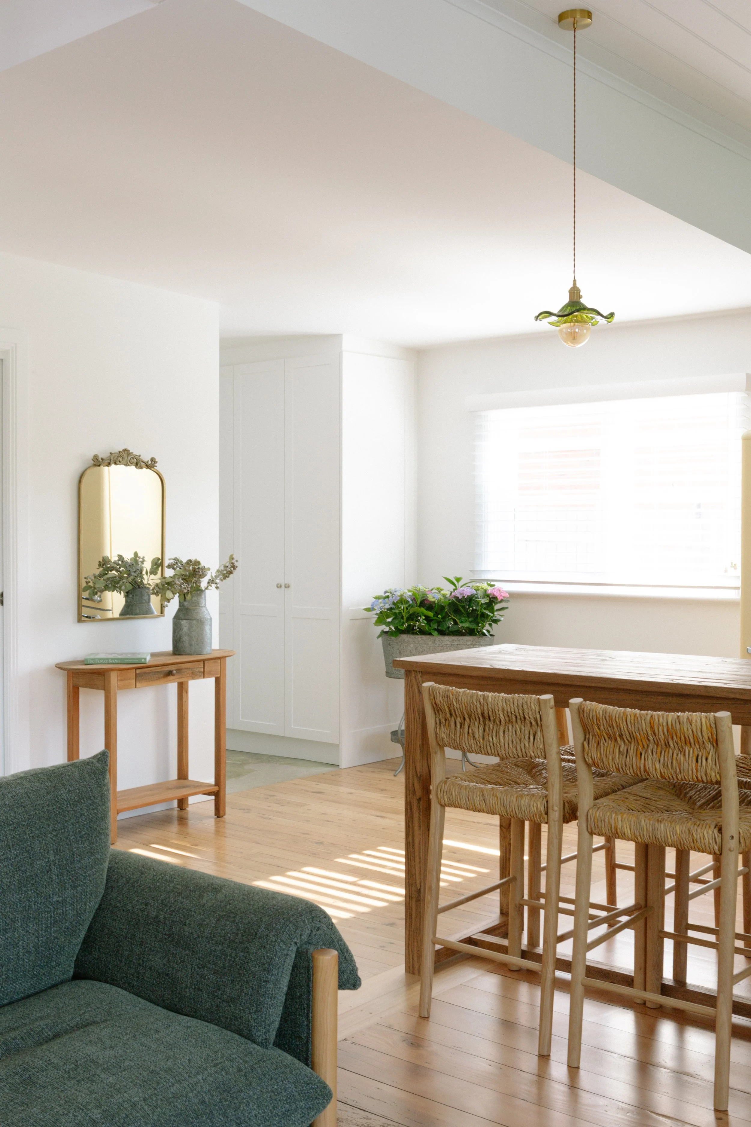 Bright living room with hardwood floors, a green armchair, a wooden dining table with woven chairs, a small wooden console table with vases and flowers, white walls, a large window with blinds, and a ceiling light fixture.