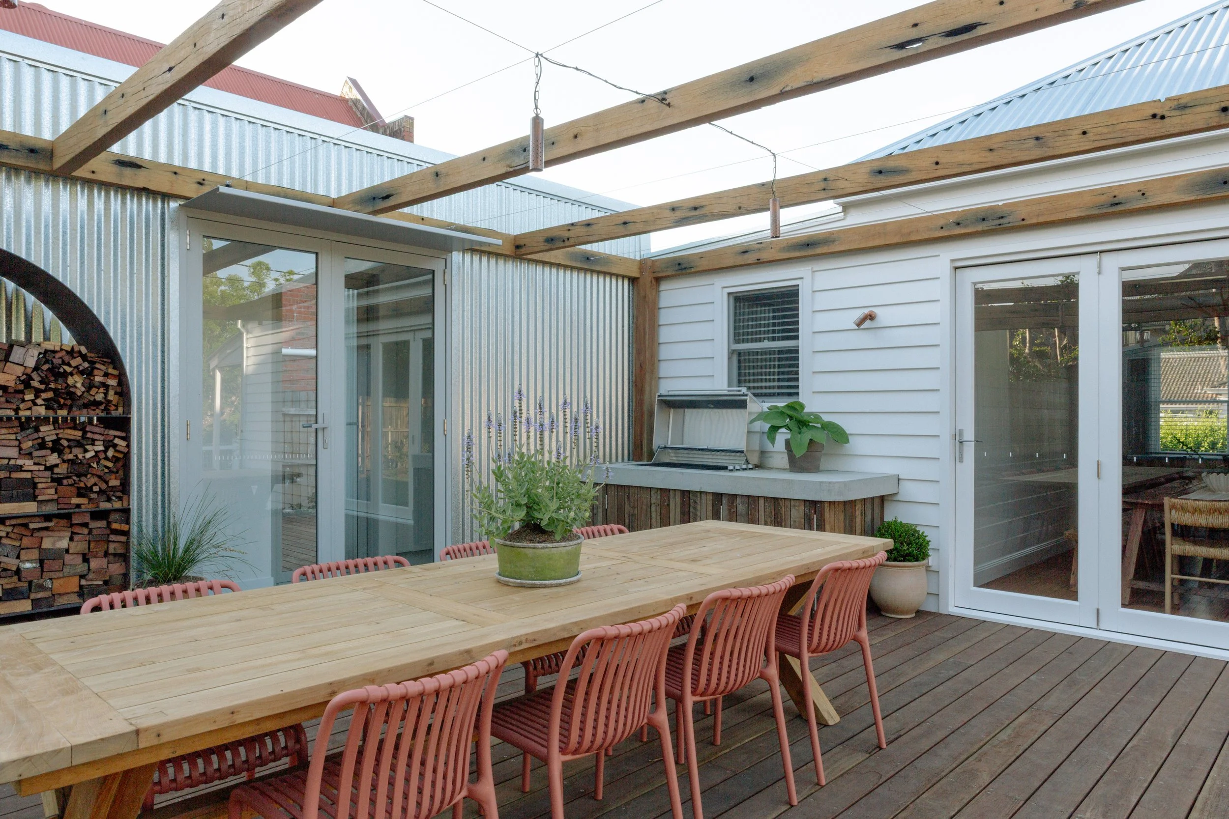 Outdoor patio with a long wooden dining table and pink chairs, potted plants, a grill, and a wood storage cabinet, with a metal and wood structure overhead and sliding glass doors leading to the interior.