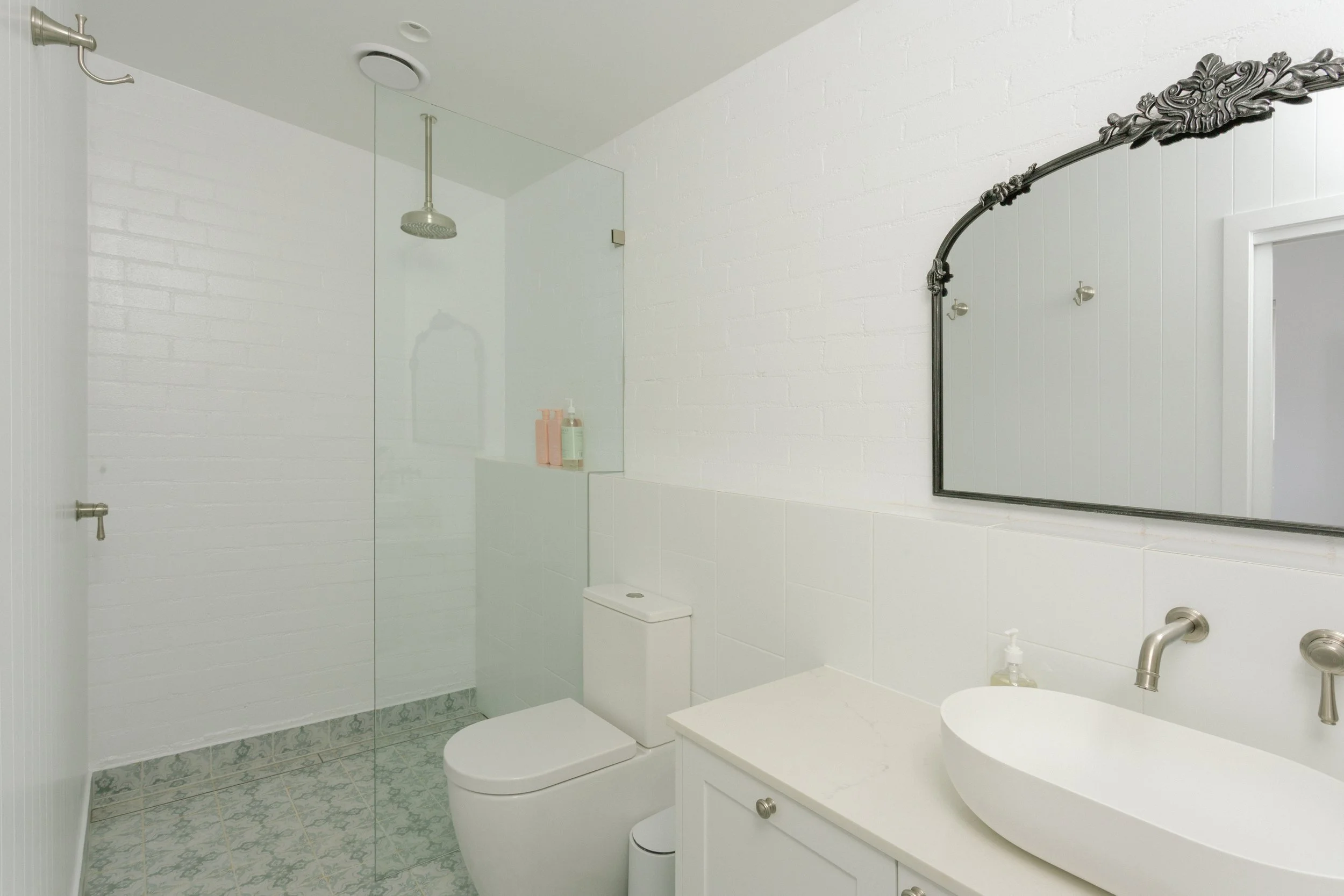Minimalist bathroom with white brick walls, a glass shower enclosure, a white toilet, a white vanity with a vessel sink, and a large mirror with ornate black frame.
