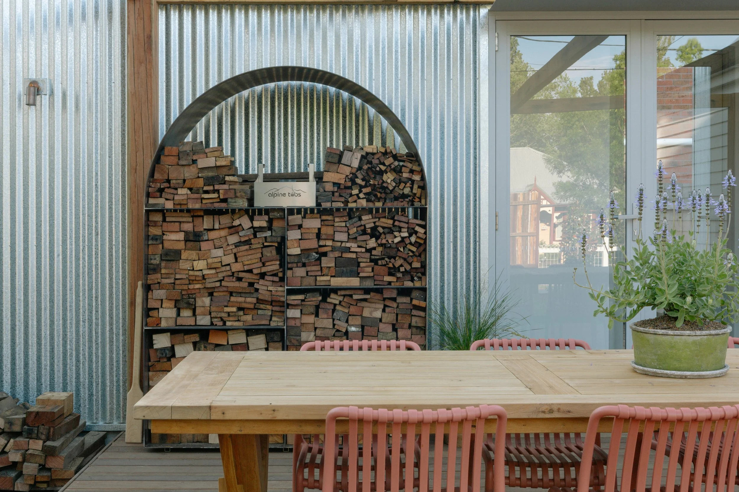 An indoor patio with stacked firewood against a corrugated metal wall, a wooden table with pink chairs, and potted plants near a large window.