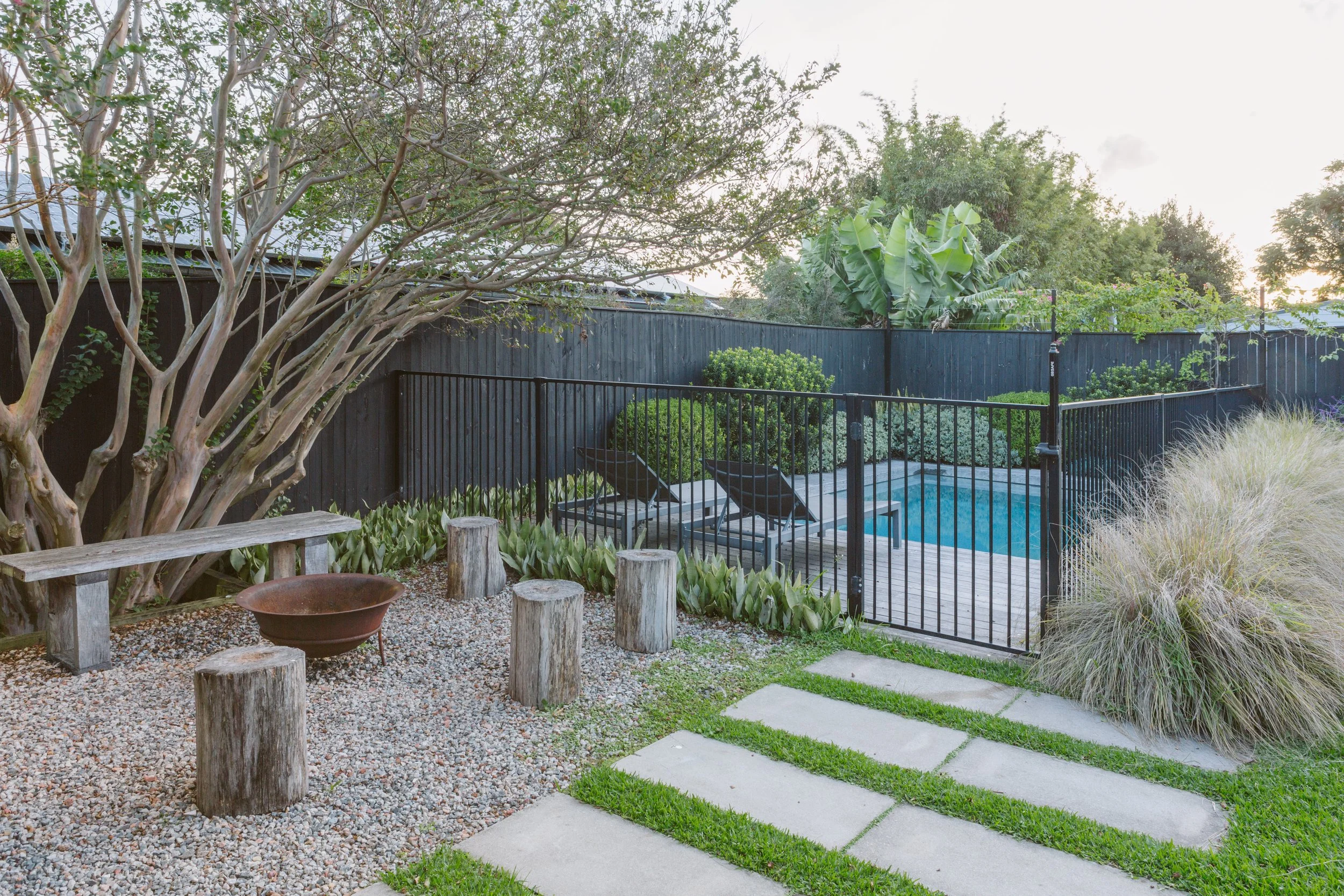Backyard featuring a small swimming pool surrounded by a black fence, lounge chairs, lush greenery, a large tree, and a decorative seating area with wooden stumps and a fire bowl.