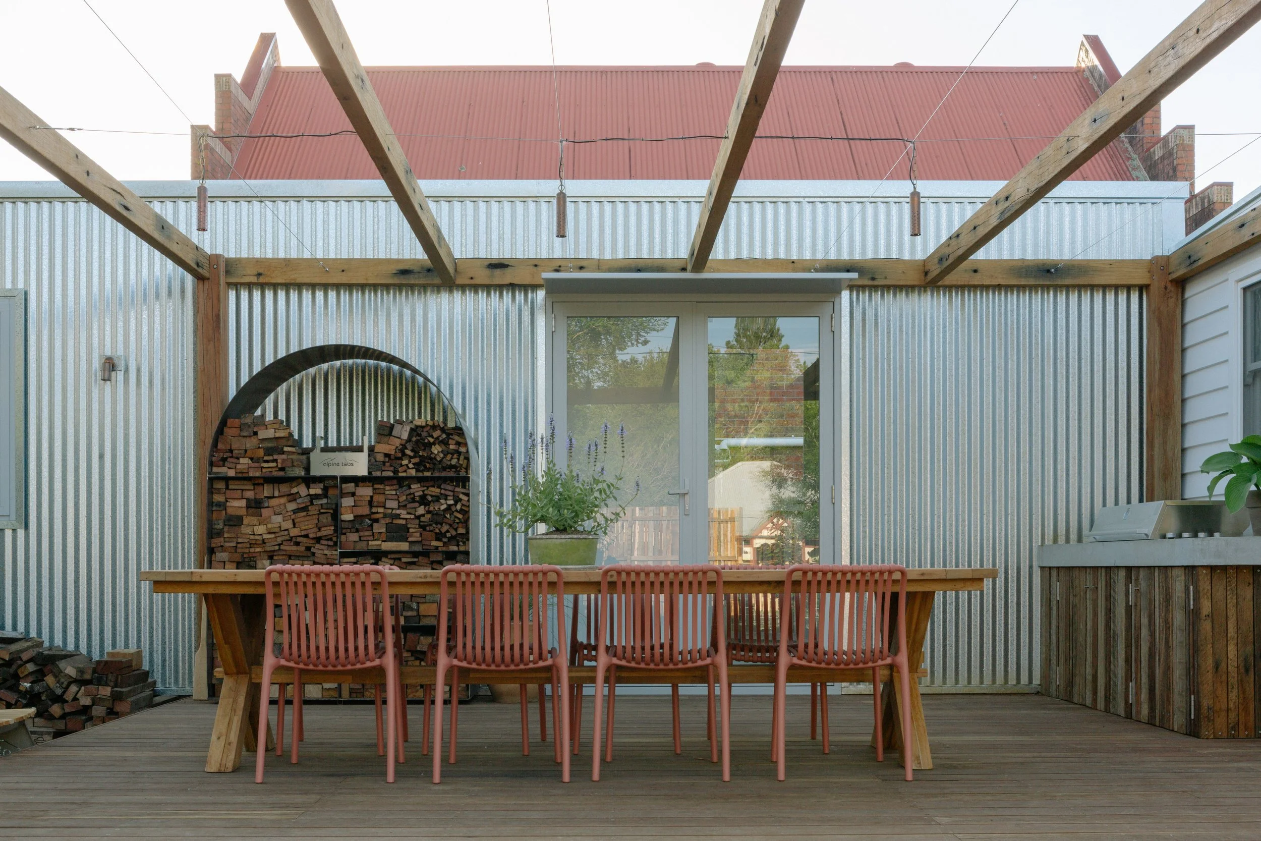 Outdoor patio with wooden table and pink chairs, a potted plant, a glass door, wood and corrugated metal walls, and a firewood storage with stacked wood, under a partly constructed pergola.