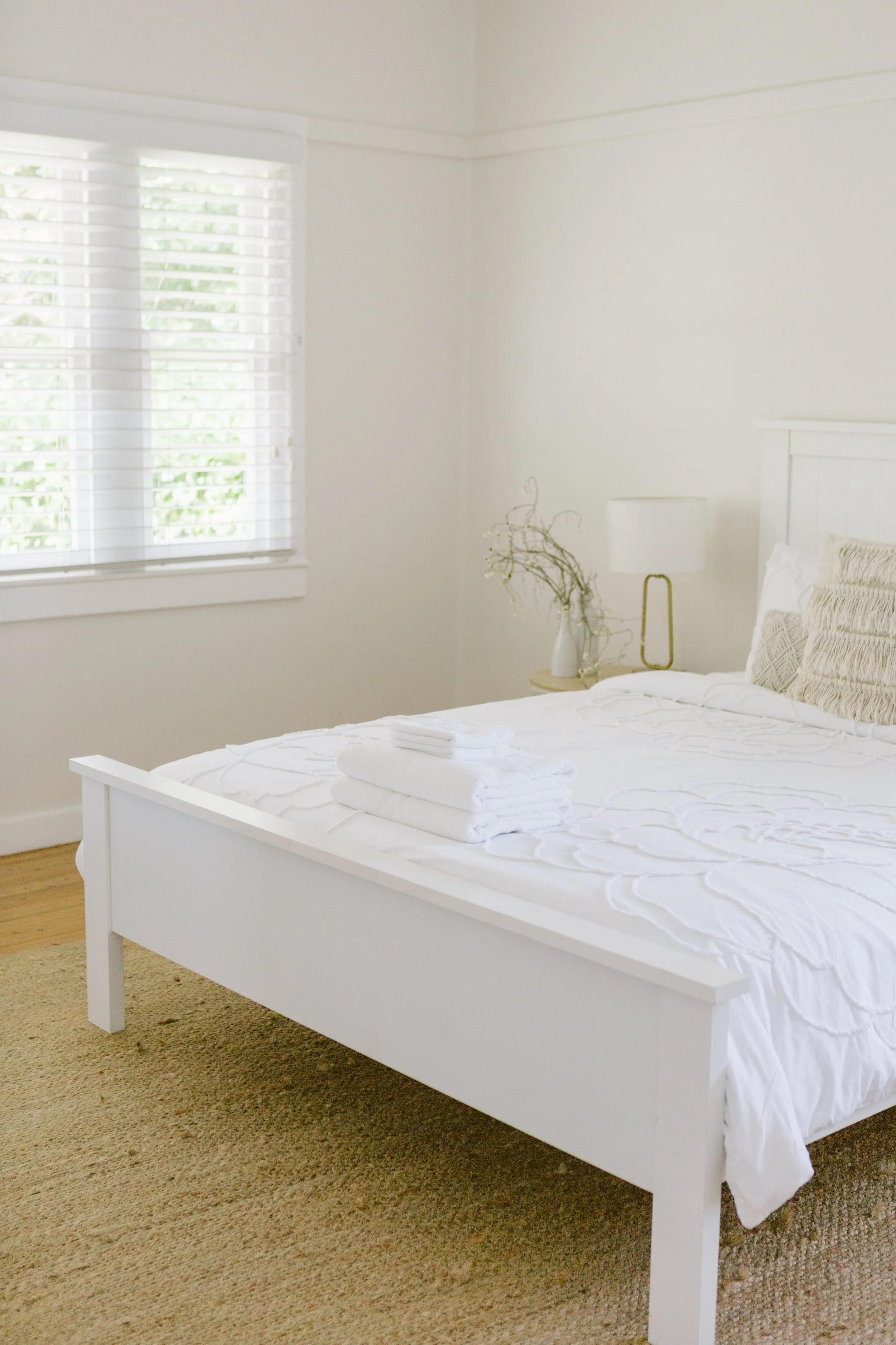 A bedroom with white walls, a white bed, folded white towels on the bed, a window with white blinds, a bedside table with a vase of dried branches, and a modern white and gold lamp.