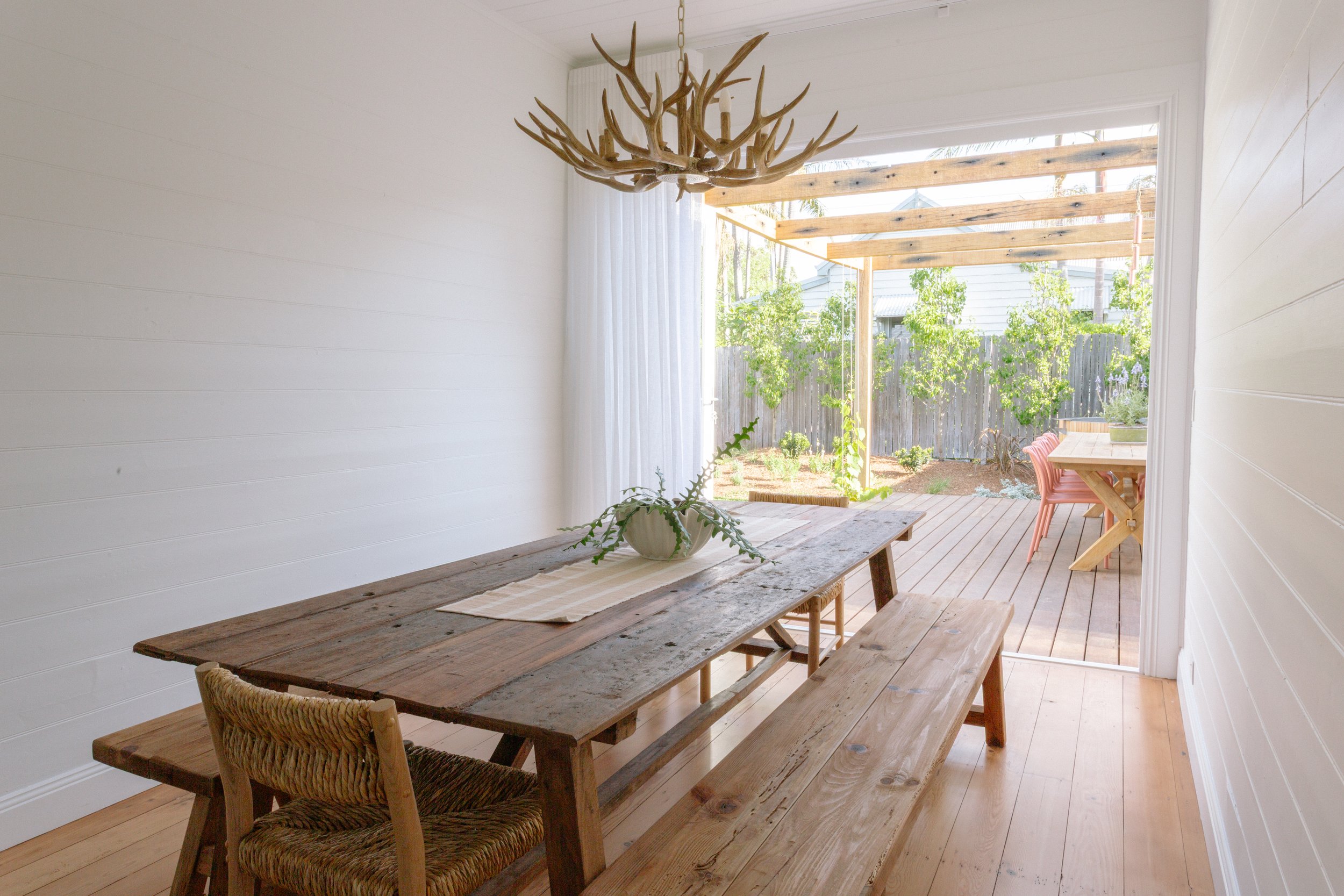 Dining room with rustic wooden table, wicker chair, bench, and a chandelier made of antlers. Opens to a backyard with a wooden pergola, garden, trees, and outdoor table with pink chairs.