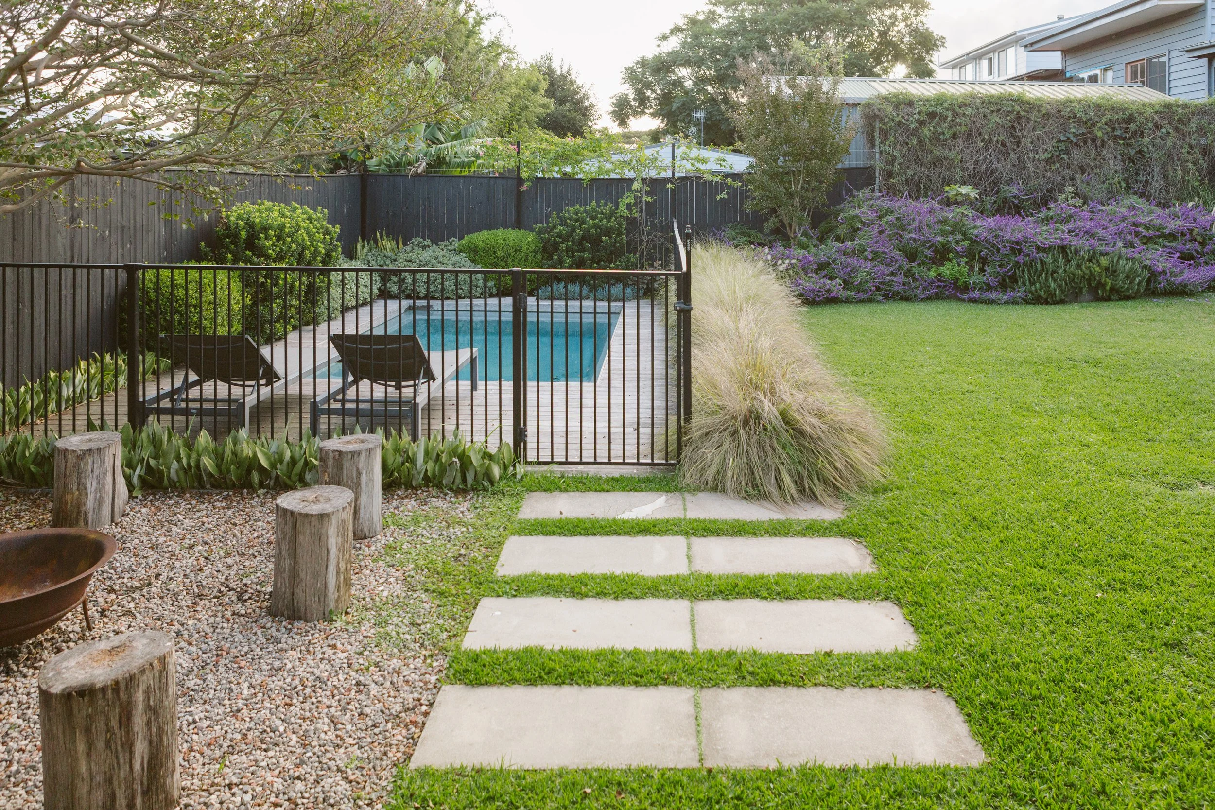 Backyard with a sitting pool, surrounded by lush green grass, trees, shrubs, and purple flowering bushes, enclosed with a black fence.