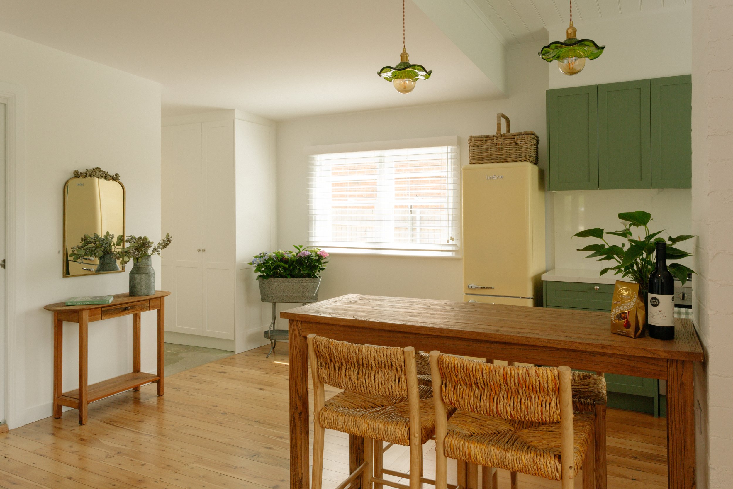 A cozy kitchen with natural wood accents, green cabinets, a vintage yellow refrigerator, a wooden dining table with woven chairs, potted plants, and decorative lighting fixtures.