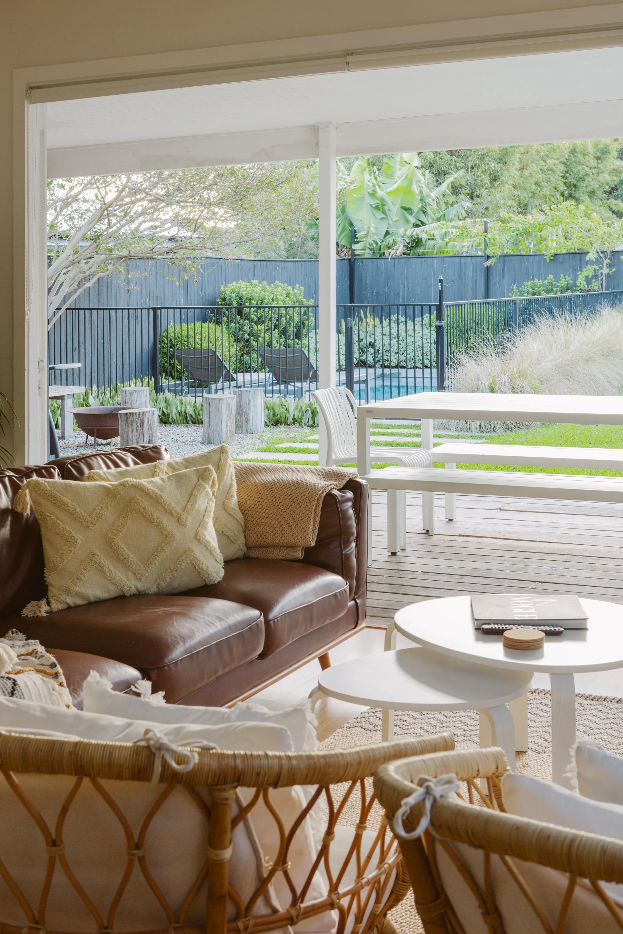 Living room with white coffee table, brown leather sofa with decorative pillows, wicker chairs, and a view of a backyard with lounge chairs, greenery, and a swimming pool.