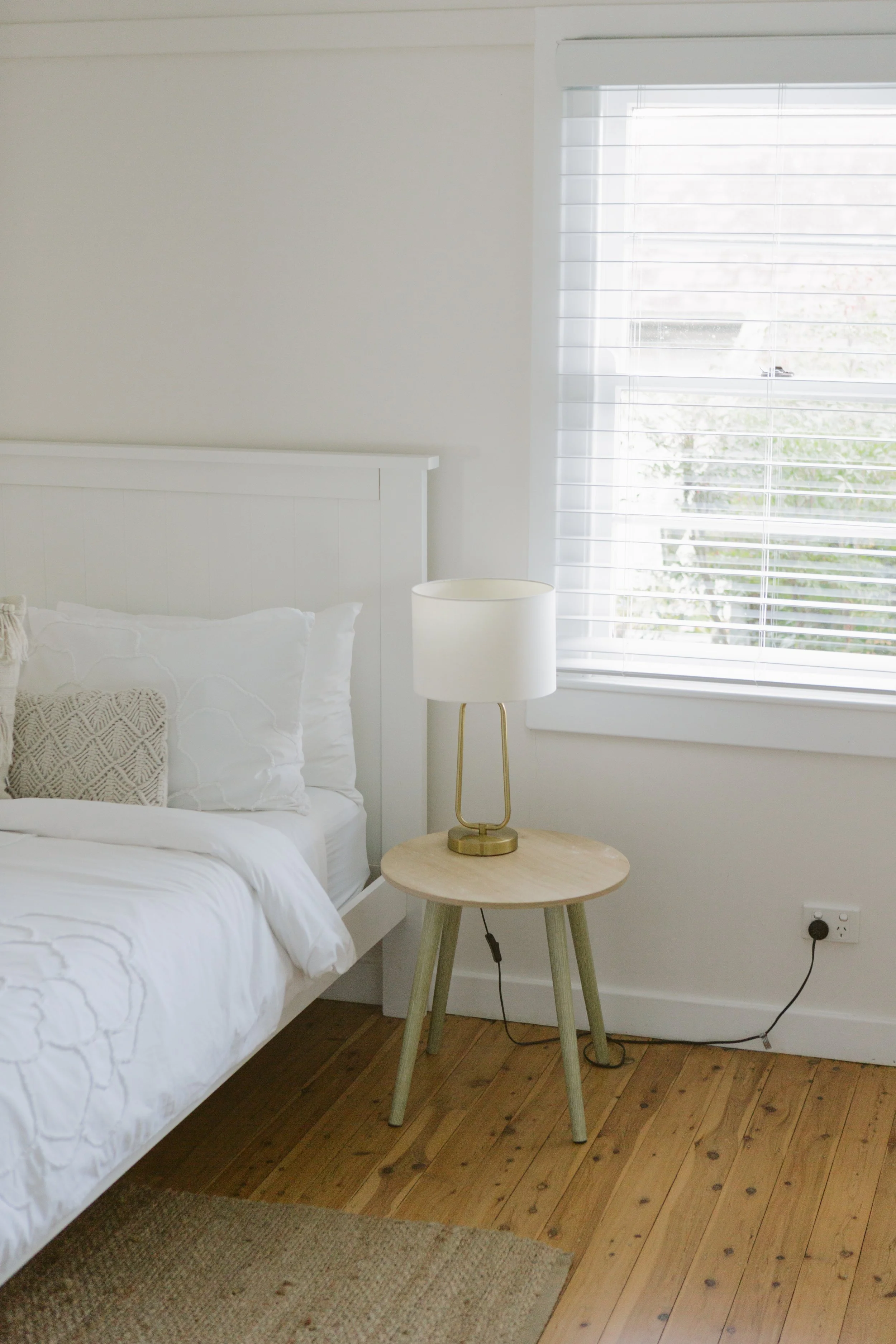A bedroom with a white bed, a small round wooden side table, a white lamp, and a window with white blinds.