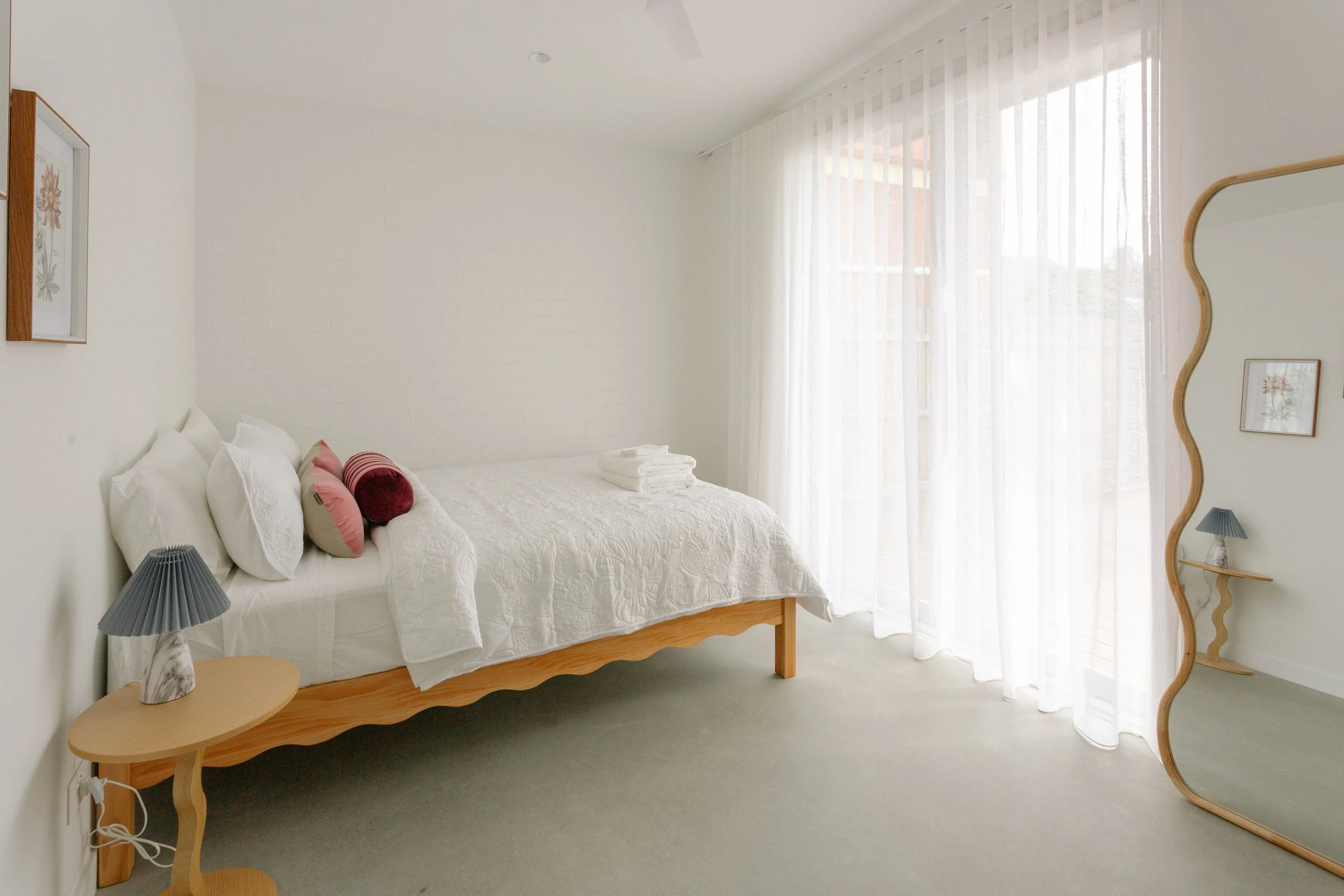 Bright, minimalistic bedroom with a bed, side table with lamp, mirrored standing mirror, sheer curtains, and a framed art piece.