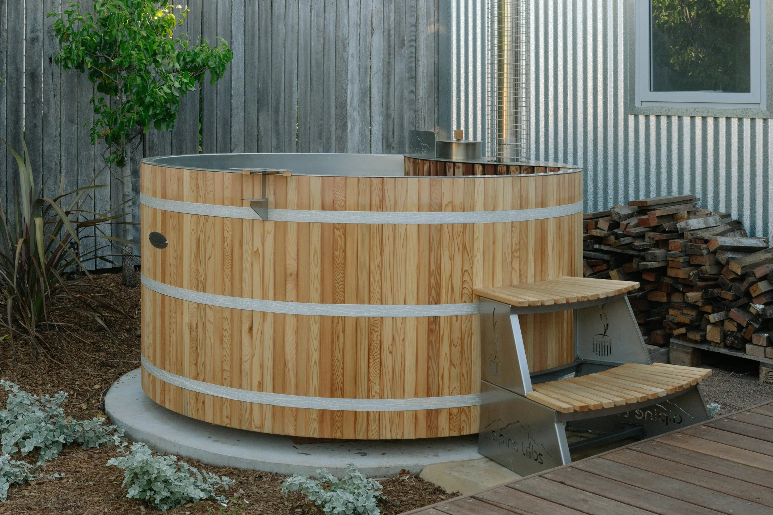 Wooden hot tub with metal interior, small wooden stairs, and a metal chimney in a backyard with a corrugated metal siding wall, a window, a tree, and a pile of firewood.