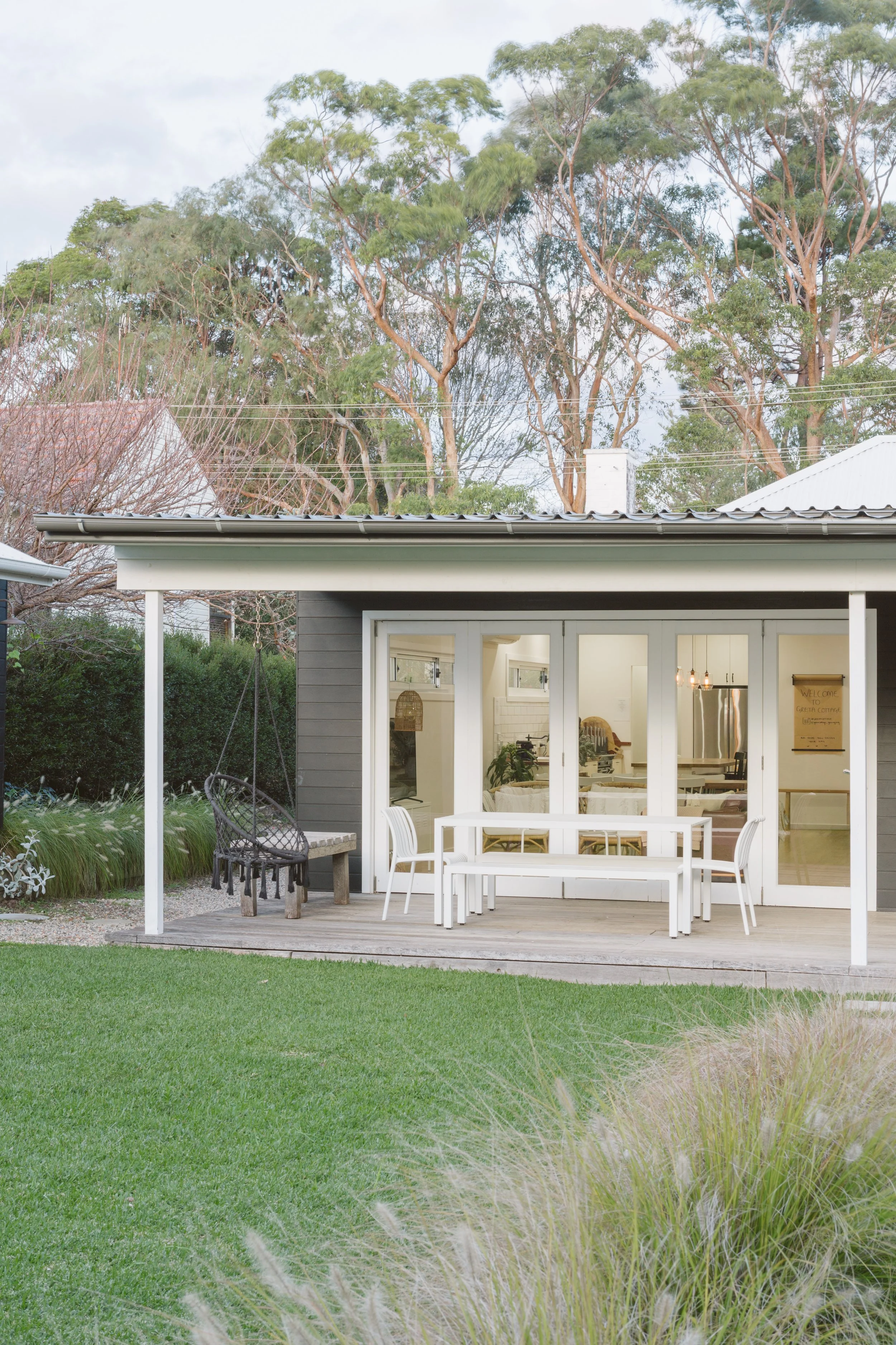 Backyard with a covered patio, white table, chairs, and a hanging swing, with green grass and trees in the background.
