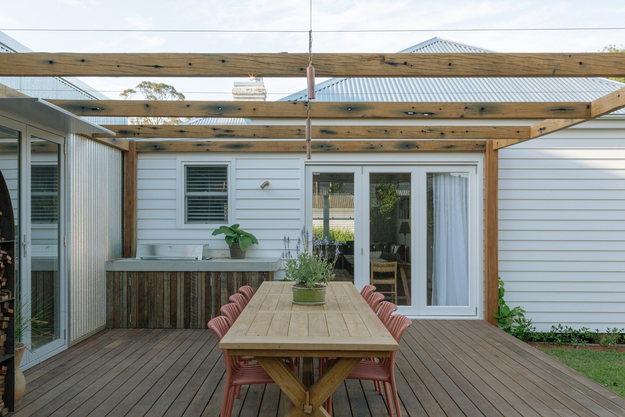 Outdoor patio with a wooden dining table and pink chairs, potted lavender plant in the center, surrounded by a partially constructed wooden pergola, sliding glass door with white curtains, and a house featuring white siding and a metal roof.