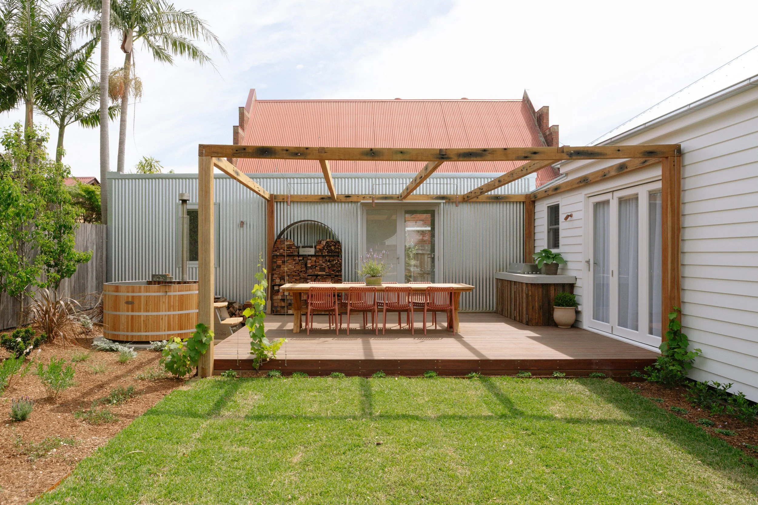 Backyard patio with wooden deck, outdoor dining table with eight chairs, a grill, potted plants, firewood stack, hot tub, and some gardening plants, with a house and trees in the background.