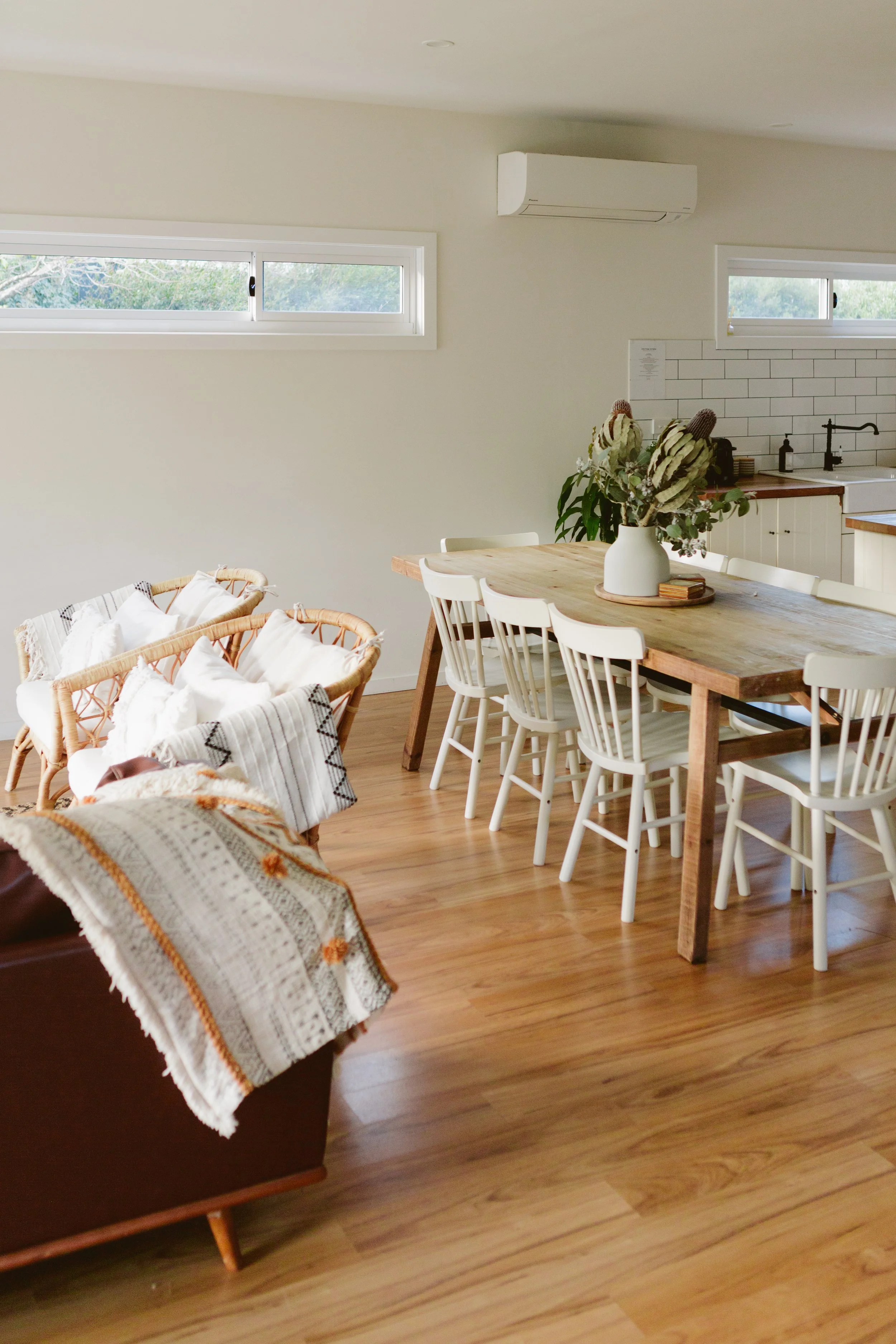 A dining area with a wooden table surrounded by white chairs, a centerpiece with dried flowers, and a kitchen in the background with white cabinetry, a black faucet, and white tiled backsplash. There are small horizontal windows and an air conditioni