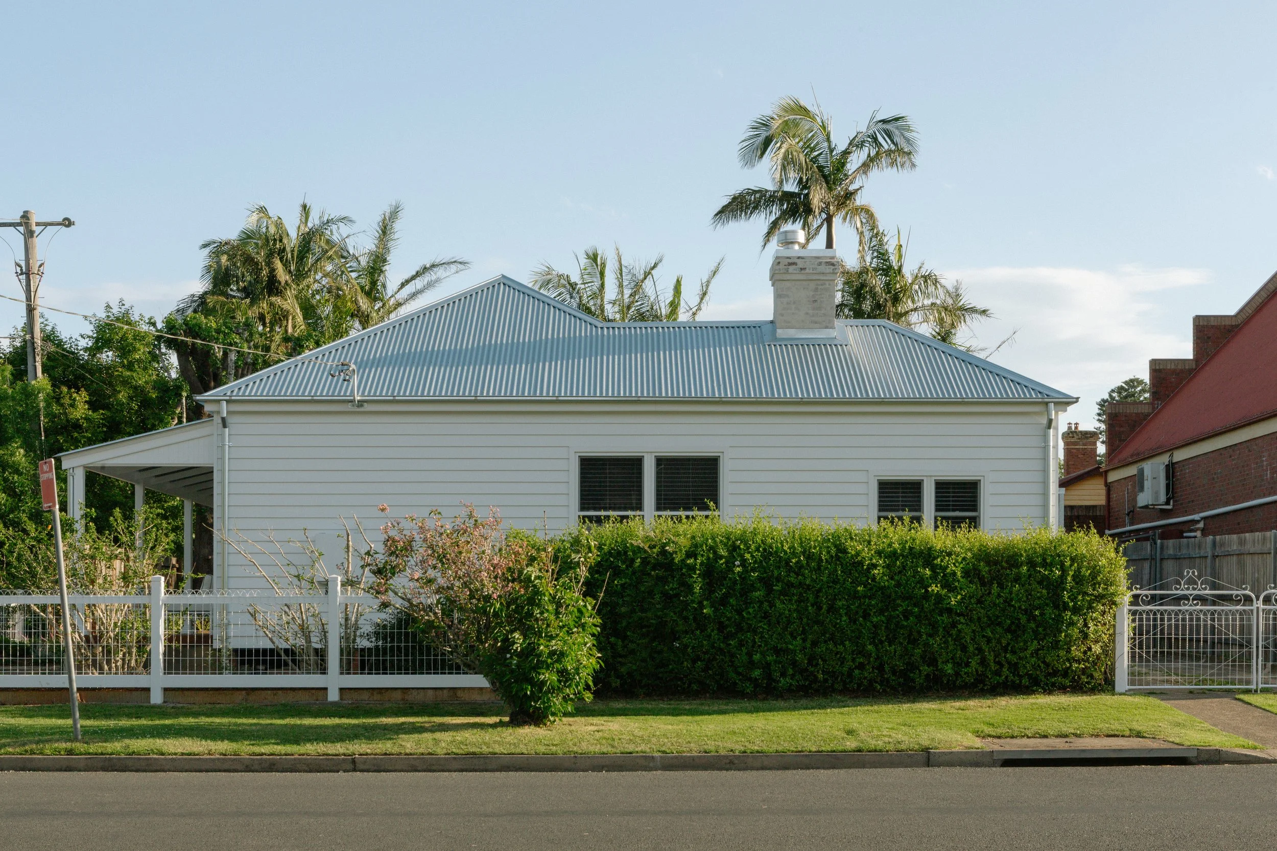 Exterior of a white house with a metal roof, surrounded by a hedge, with a white fence and sidewalk in front, and palm trees in the background.