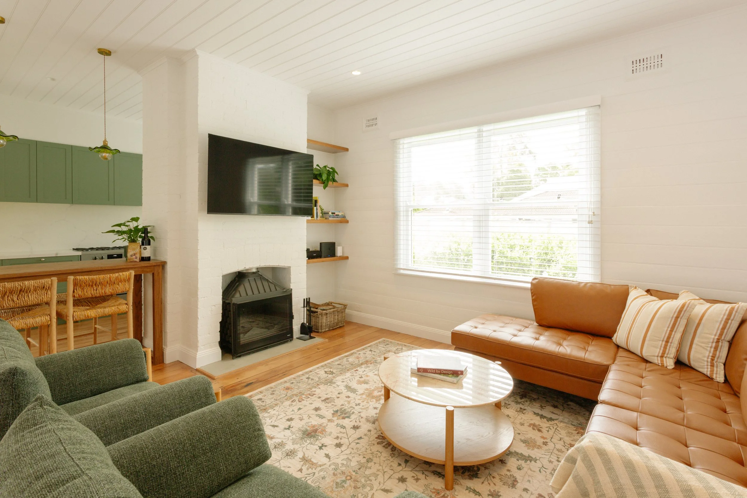 Living room with a brown leather sofa, green armchairs, a round coffee table, a white brick fireplace, a large window with blinds, and a mounted flat-screen TV.