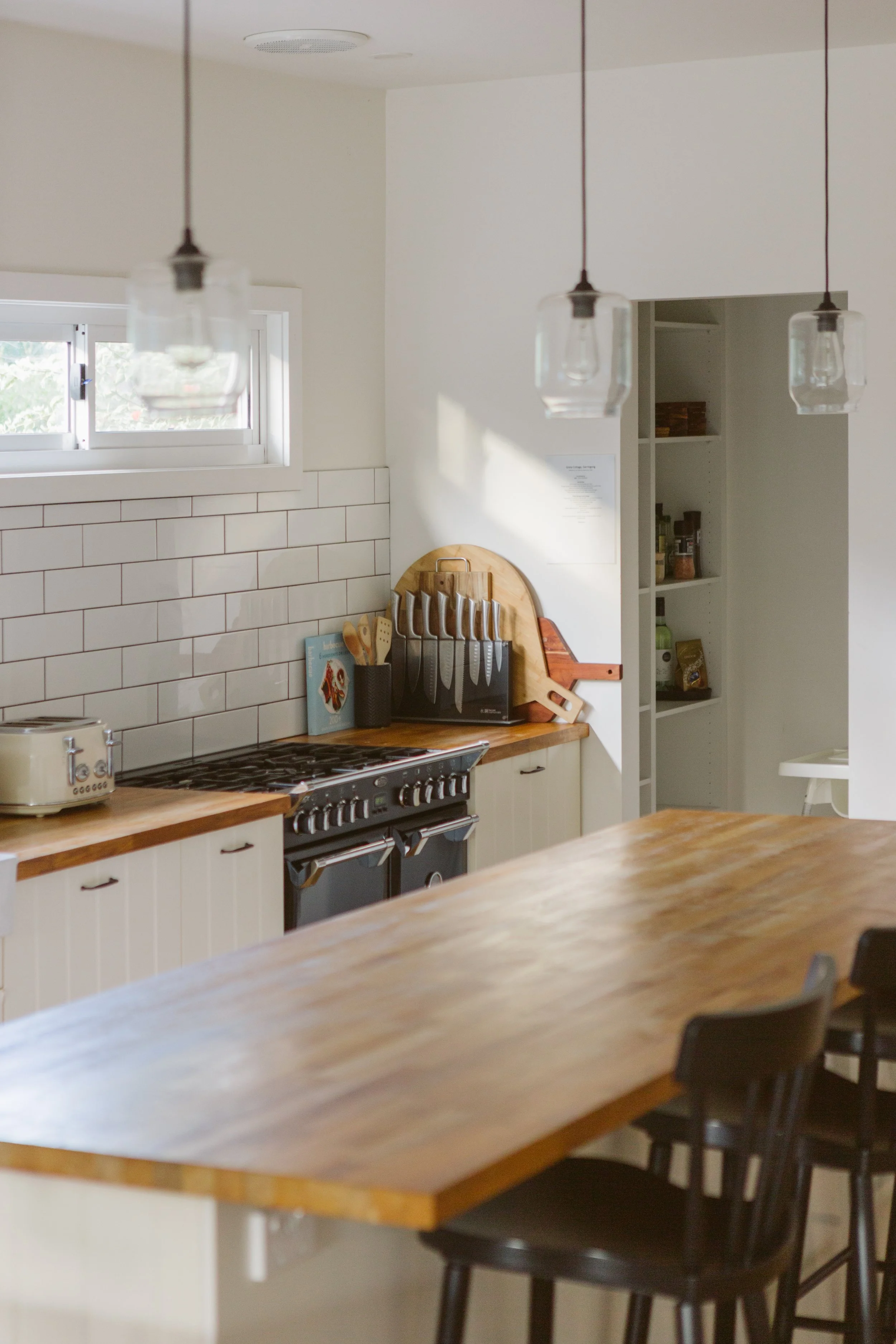 A modern kitchen with a large wooden island, black chairs, a window with sunlight, a stove, and a cutting board with knives on a wooden stand.