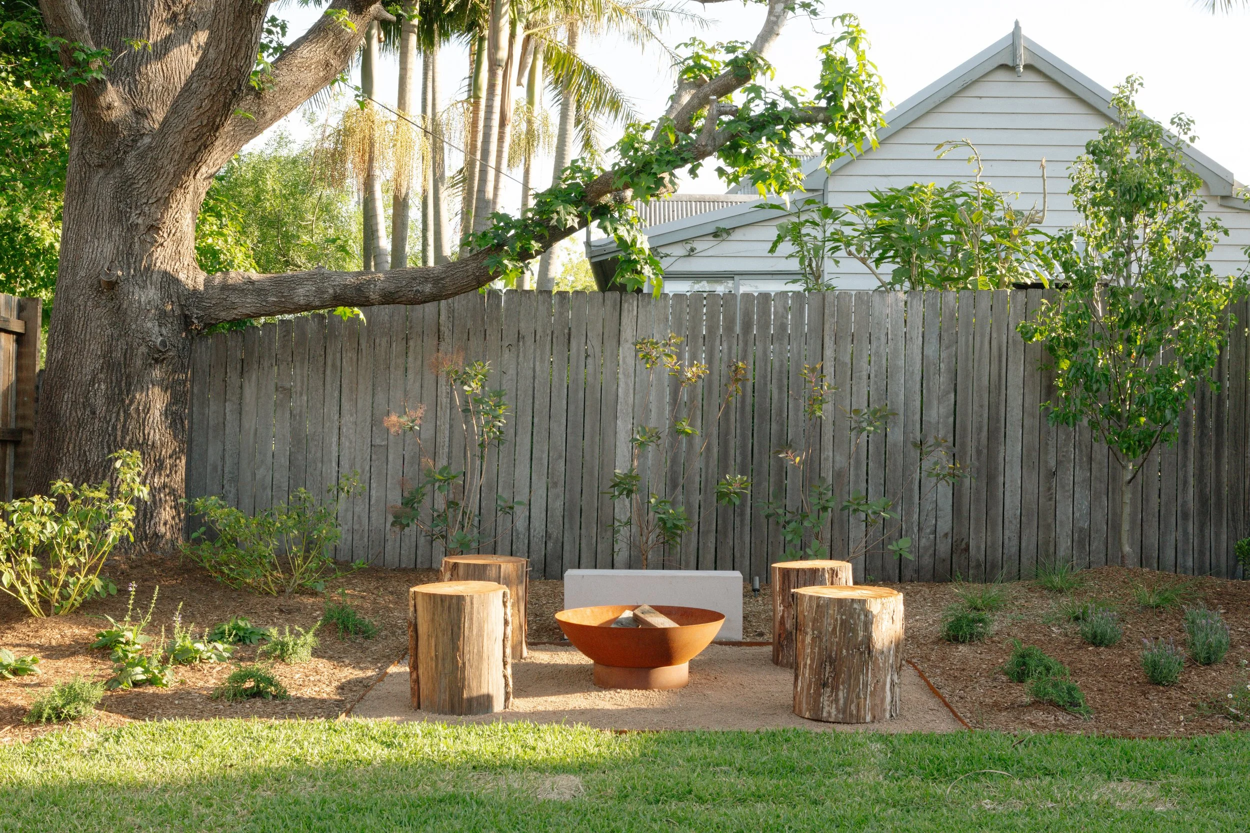 Backyard with a large tree, a wooden fence, and a garden area featuring shrubs, small plants, and five tree stump seats surrounding a fire pit, with a whitehouse partially visible behind the fence.