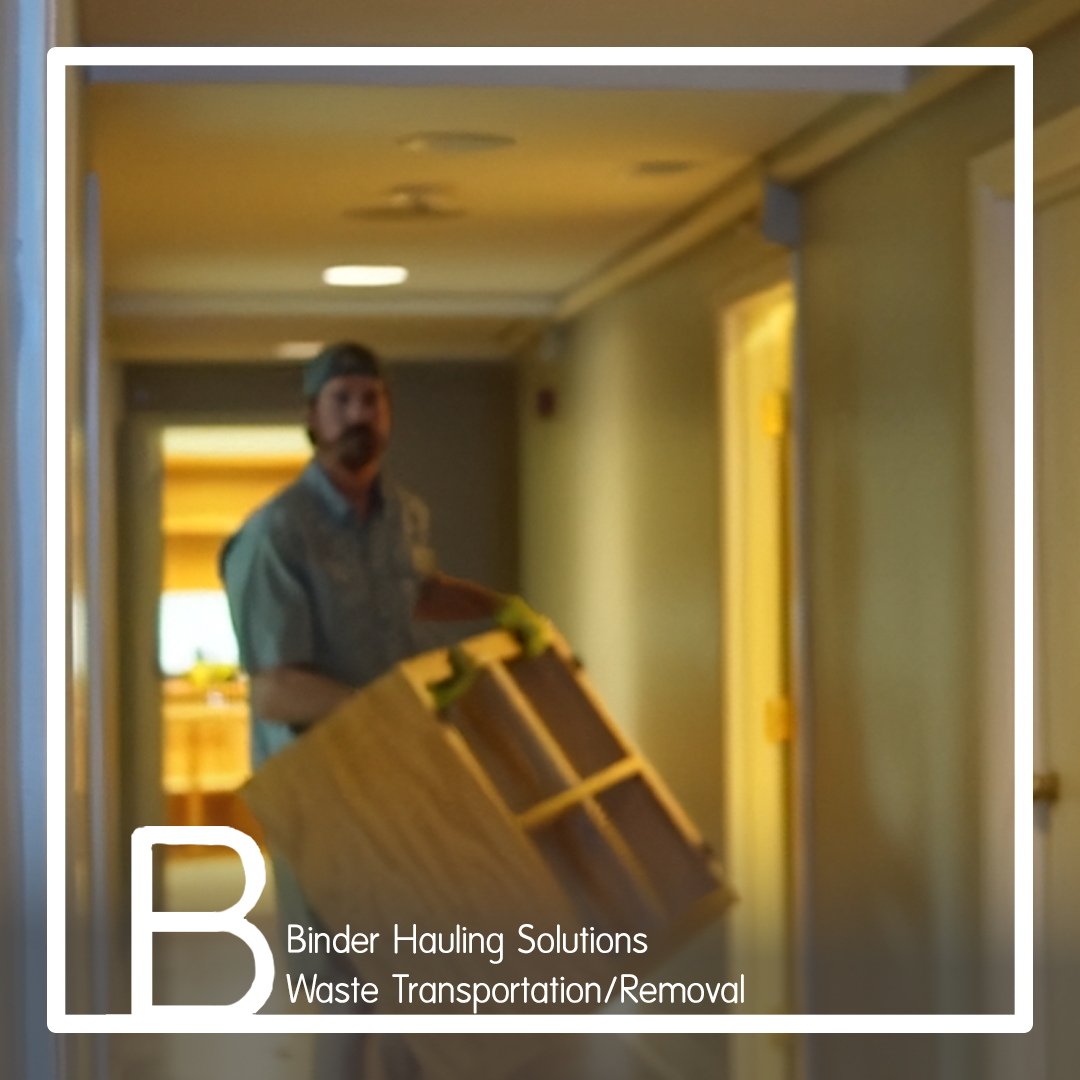 A man wearing a cap and gloves is hauling a large cardboard box in a hallway of a building.