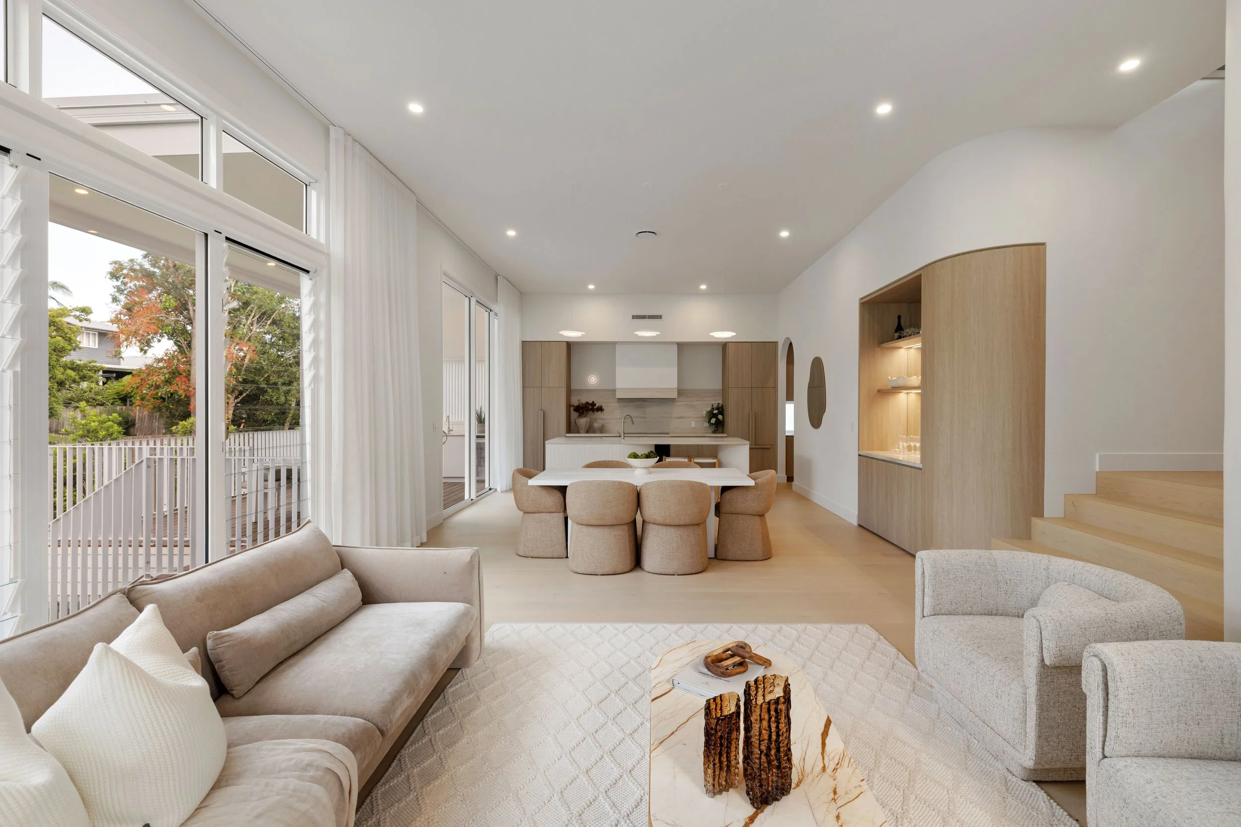 Open-concept living and dining area with neutral tones, large windows, and a view of a backyard with trees.