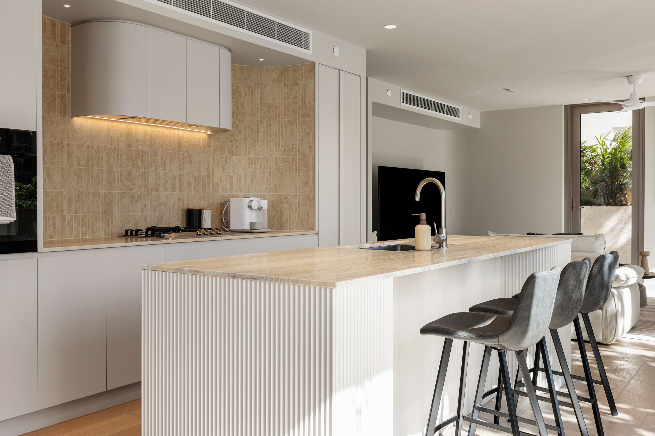 Modern kitchen with white cabinets, beige tiled backsplash, and a white island with seating. There is a coffee maker, stove, and wall-mounted cabinets. The space is bright with natural light from large windows.