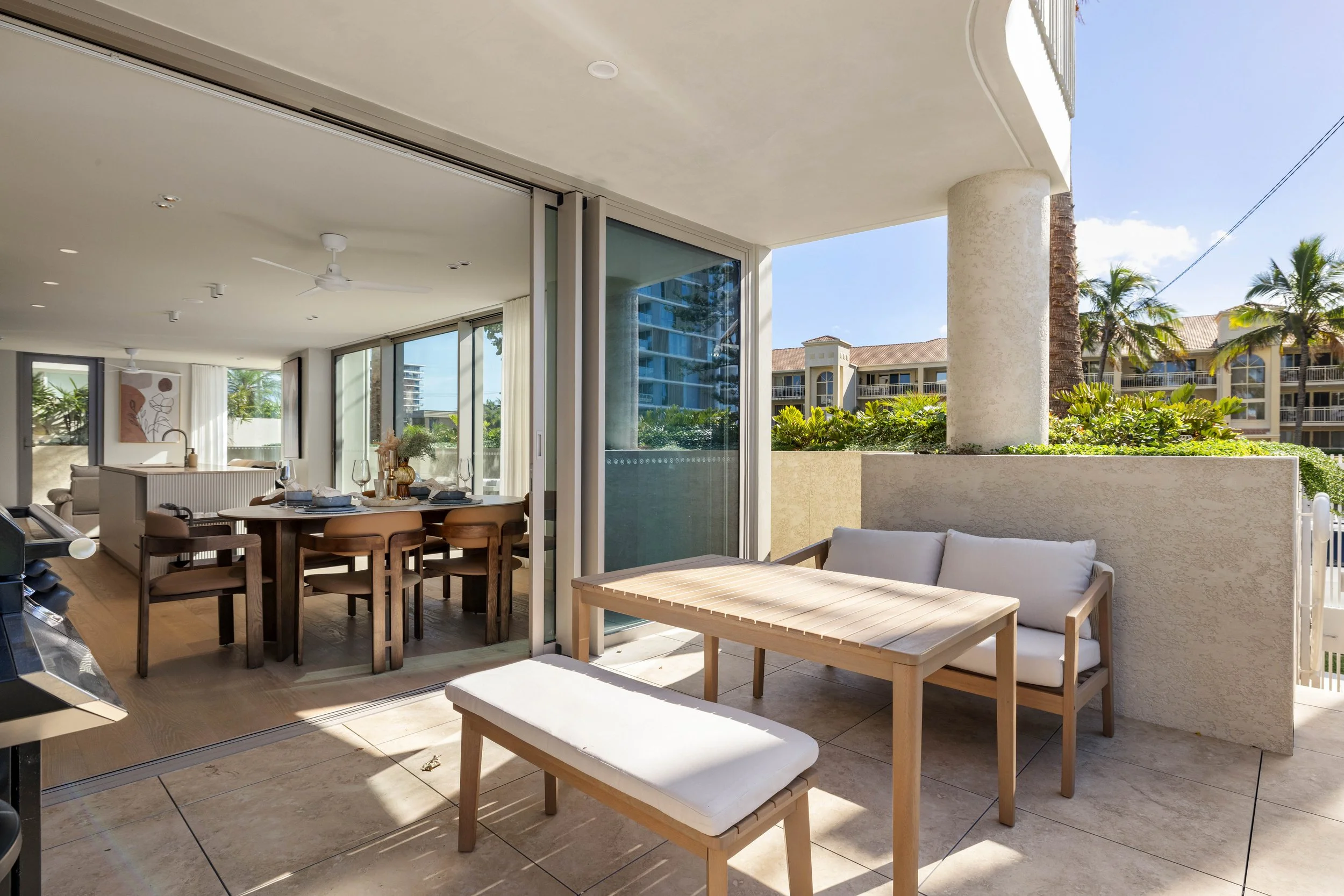 Balcony with wooden furniture, including a small table, a cushioned bench, and a chair, overlooking palm trees and residential buildings in a sunny, tropical setting, with an indoor dining and living area visible through sliding glass doors.