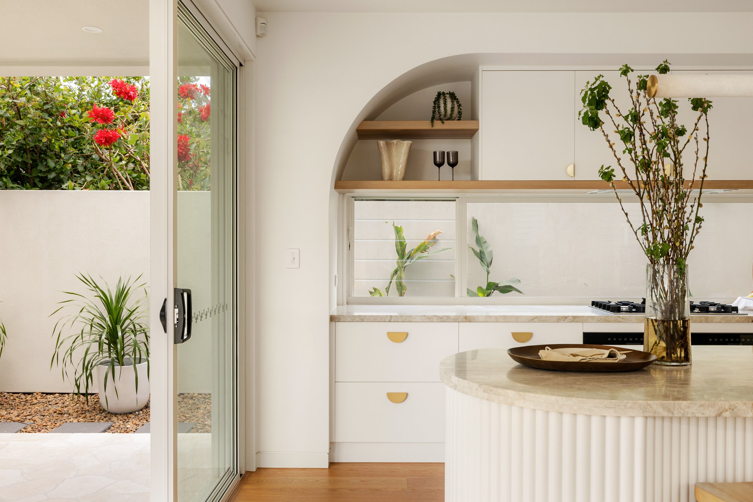 Kitchen with white cabinets, a marble countertop, and a glass sliding door leading to an outdoor area with flowering bushes and potted plants. A large vase with branches and greenery is on the island.