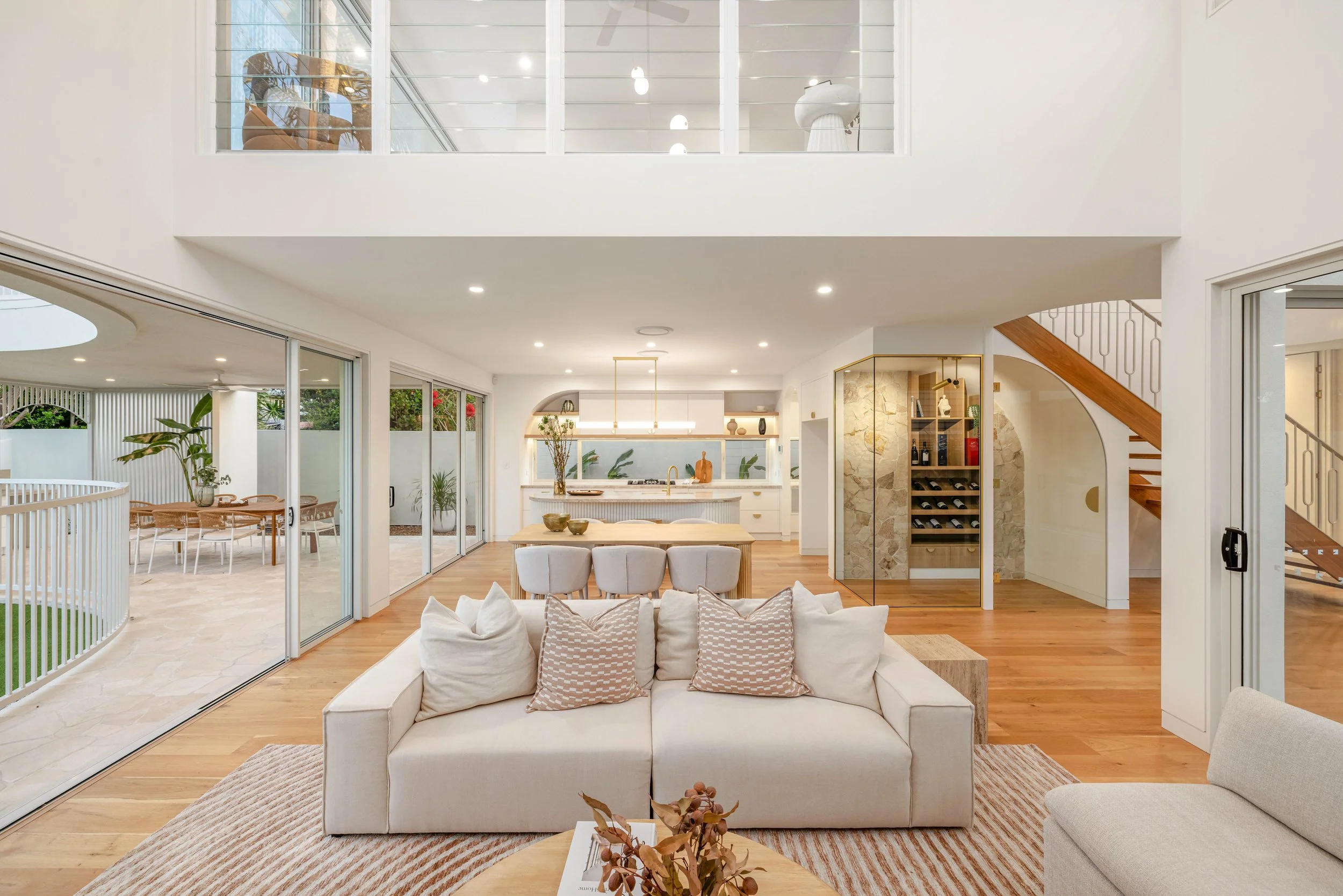 Bright, modern living room with white sofa, beige throw pillows, hardwood floors, and large glass sliding doors leading to outdoor patio with dining area and greenery.