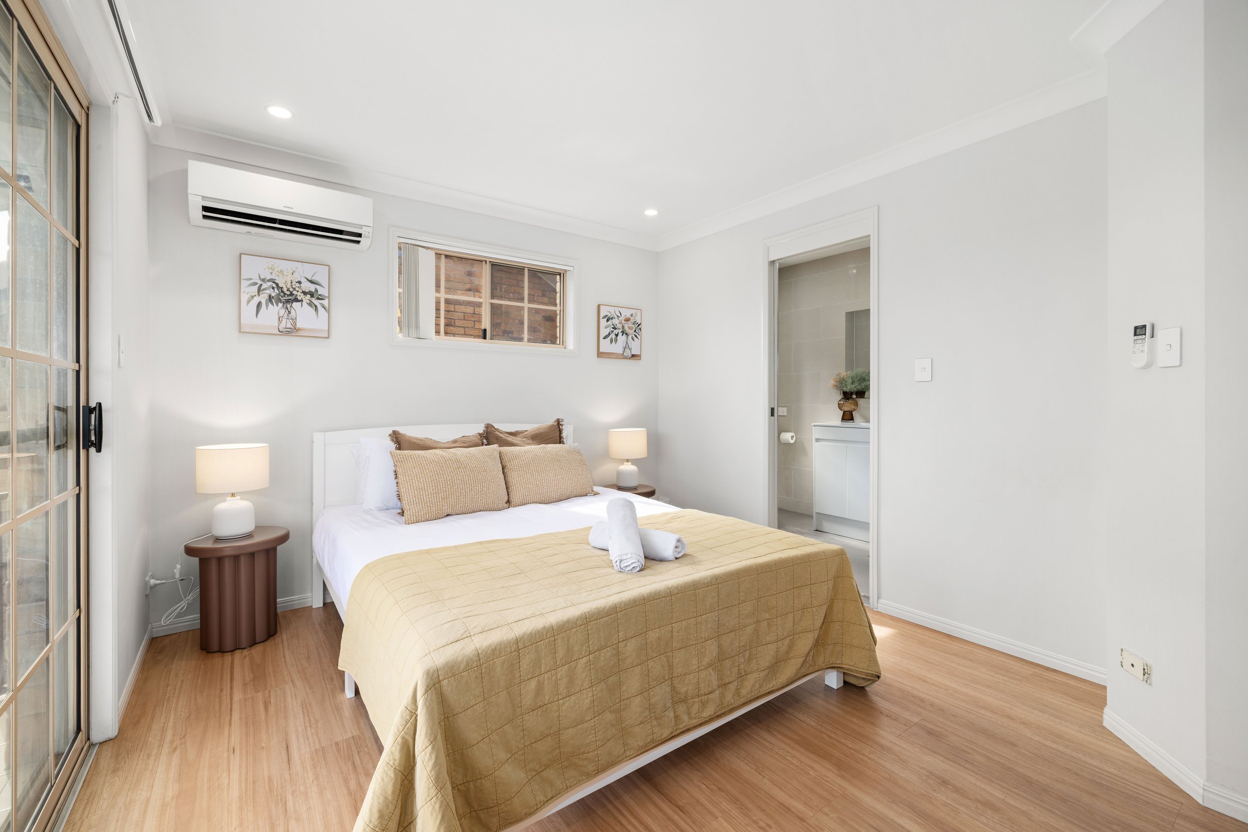 A bedroom with a white bed, beige quilt, and beige pillows. Two small wooden nightstands with lamps, artwork on the walls, and a bathroom visible through an open door.