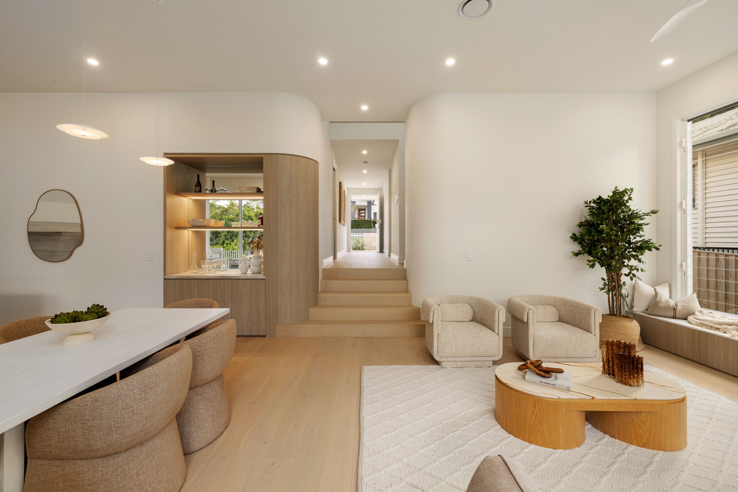 Bright modern living room with white walls, beige armchairs, a built-in window seat with cushions, wooden coffee table, potted plant, dining area with a white table and cushioned chairs, decorative mirror on the wall, and a view of the outdoors throu