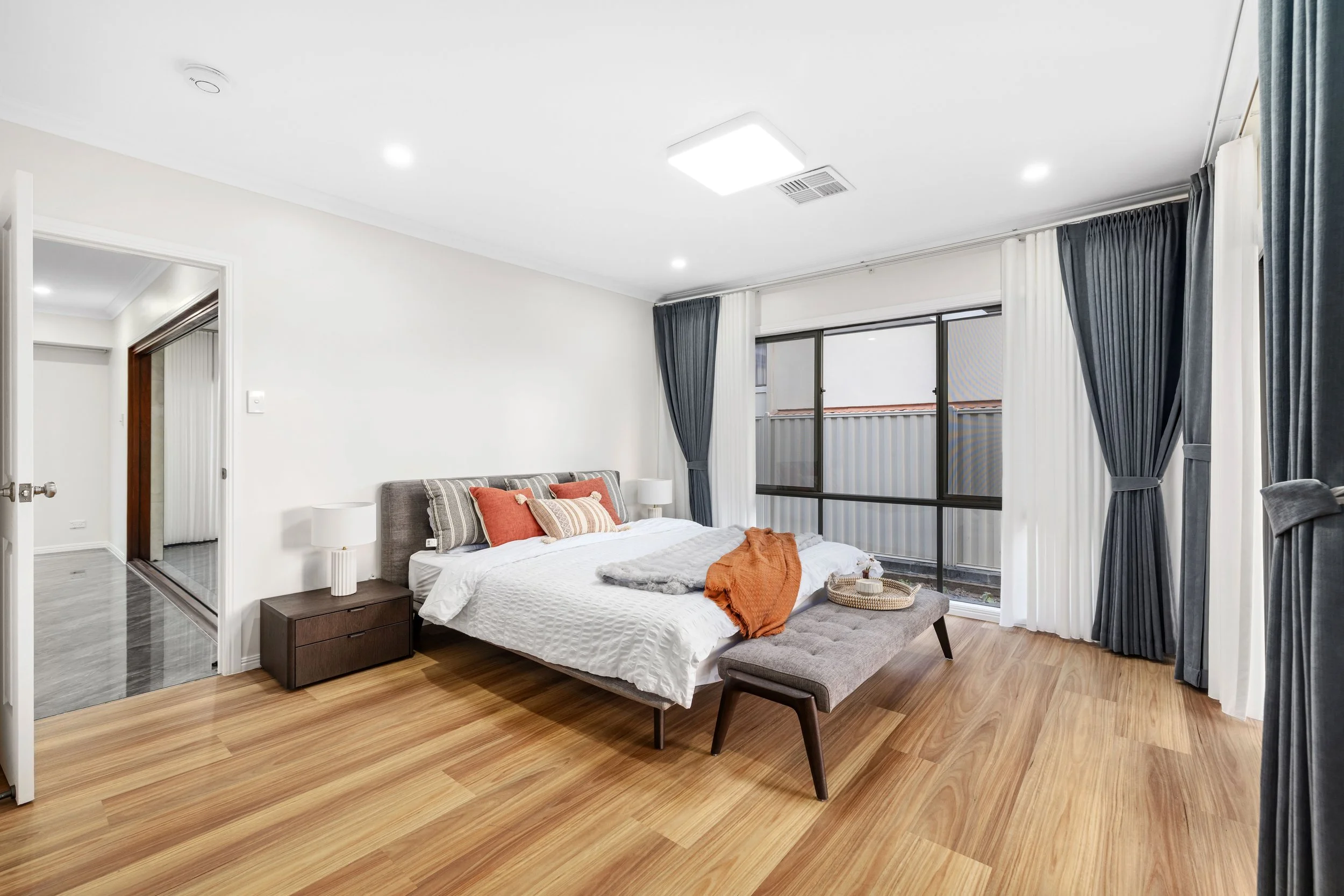 Modern bedroom with hardwood floor, gray upholstered bed with orange and beige pillows, gray bench at foot of bed, white and black curtains, large window, and minimal décor.