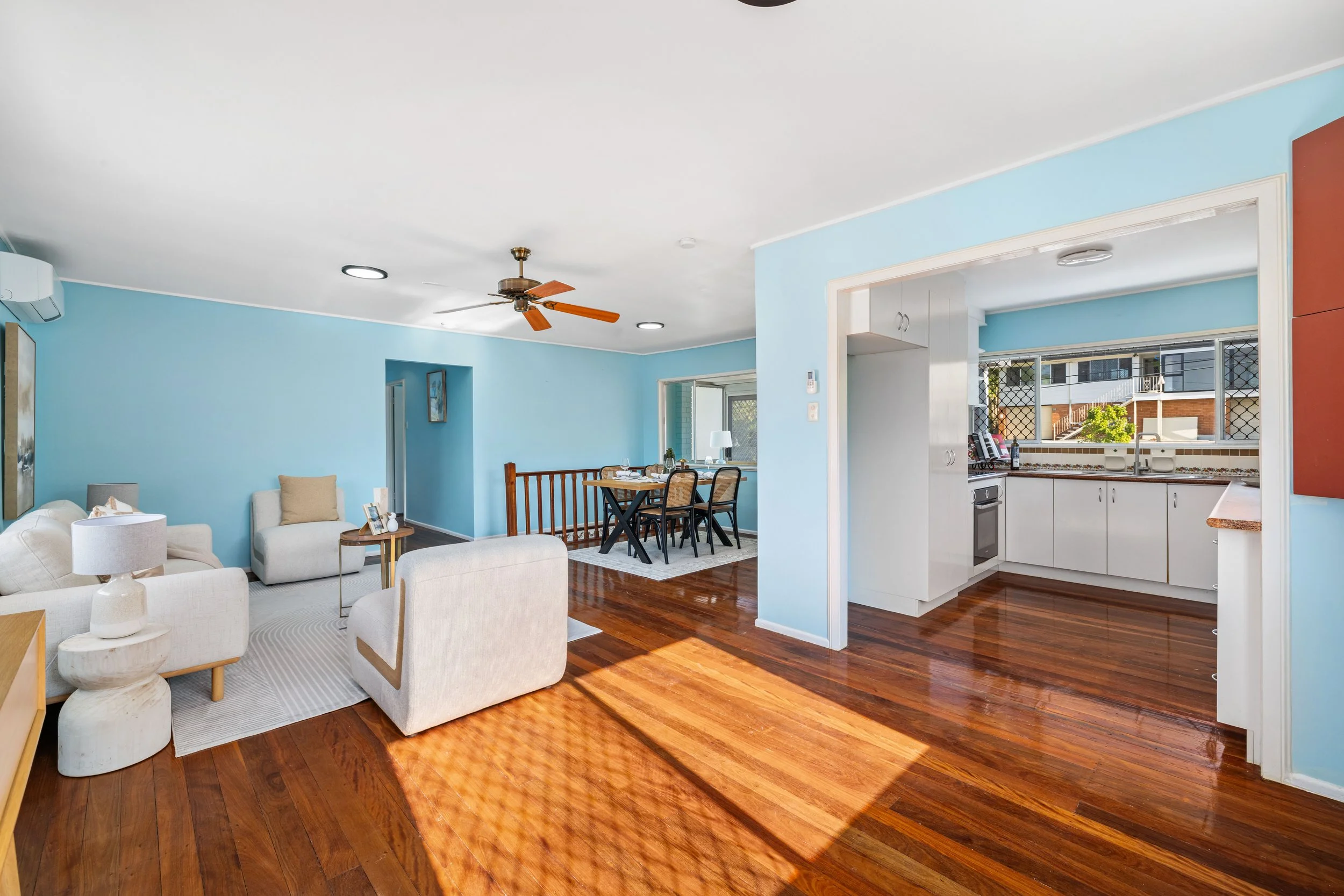 Open-concept living room and kitchen with light blue walls, wooden flooring, beige furniture, and a dining area with a window