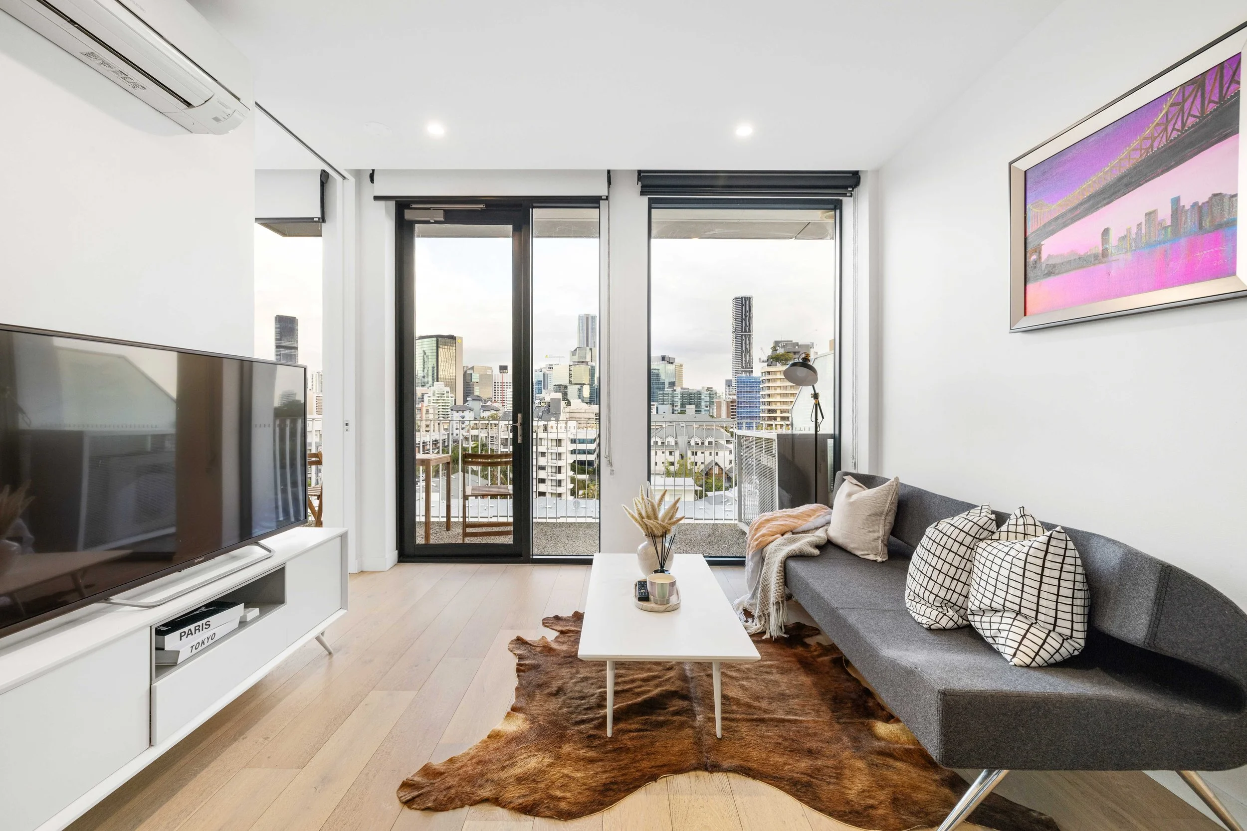 Living room with a gray sofa adorned with patterned cushions and throw blanket, white coffee table, cowhide rug, and a large flat-screen TV on a white stand. Floor-to-ceiling windows and sliding door leading to a balcony with city skyline view, inclu