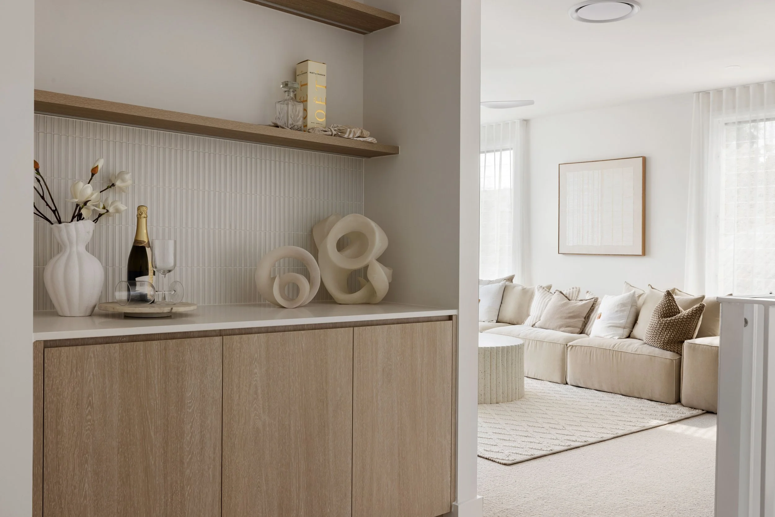 Neutral-colored living room with beige sofa, white curtains, and abstract wall art. Partial view of a wooden sideboard with decorative ceramic shapes, a vase with white flowers, and a bottle of champagne with wine glasses.