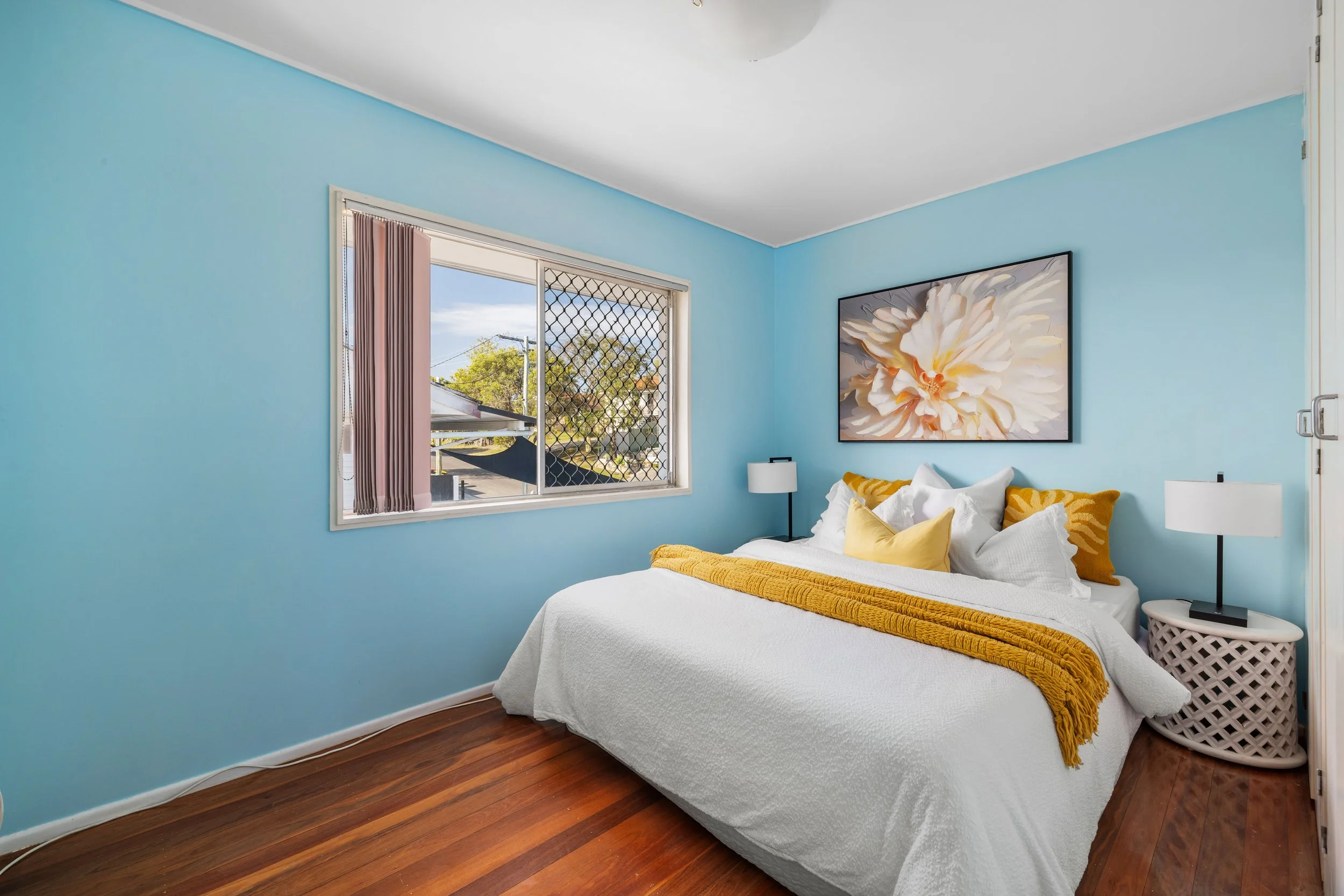Bedroom with light blue walls, a large window with a view of a street and trees, a bed with a white cover, yellow and white pillows, and a yellow throw blanket, hanging floral artwork, and two white bedside lamps.