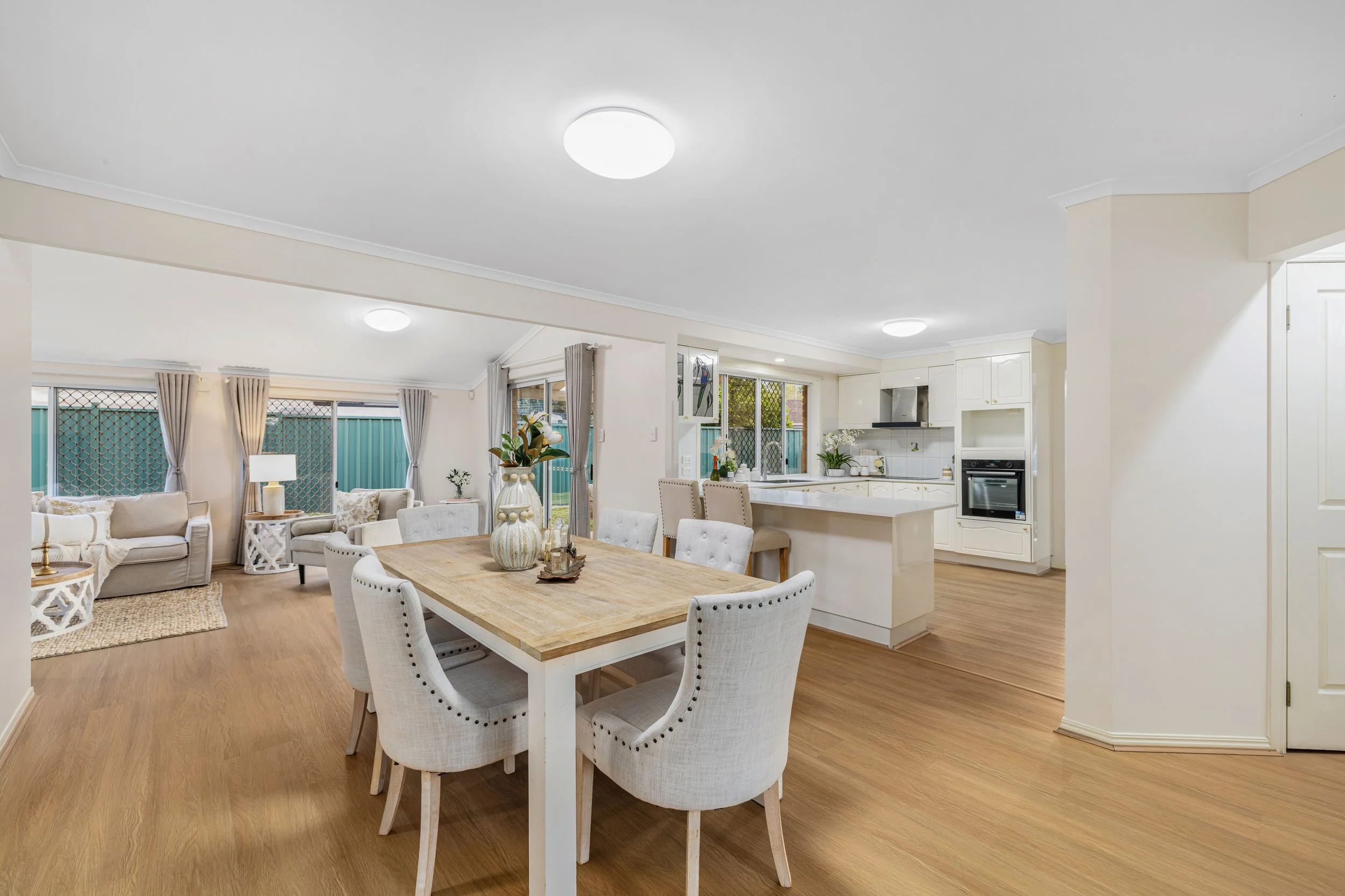 Open-concept living and dining area with light-colored furniture, hardwood floors, and a white kitchen with an island.